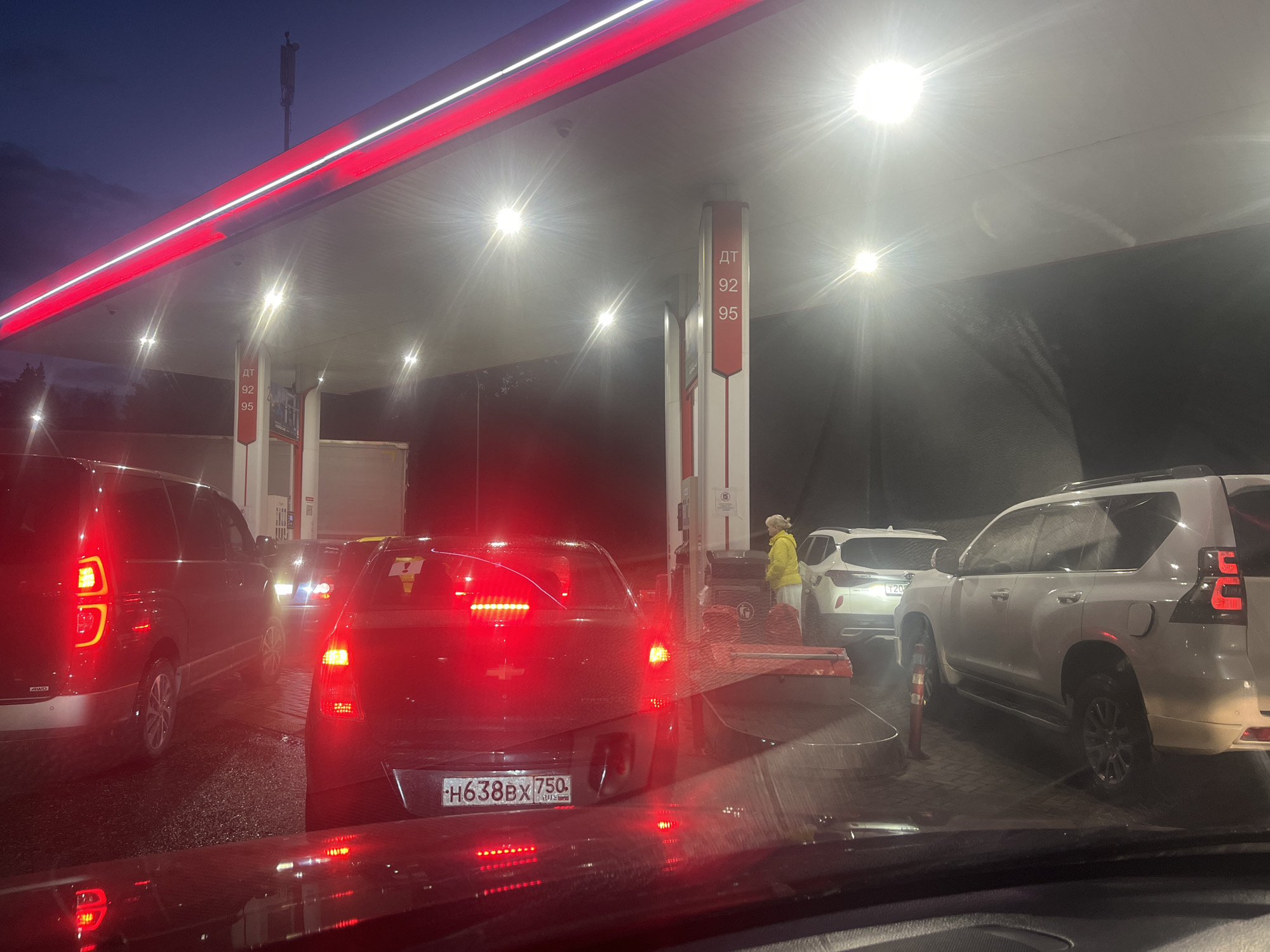 At a gas station at dusk, a queue of vehicles is observed. In the foreground, a dark-colored SUV is visible from the rear, with its red taillights illuminated. To its left, a black minivan is parked, its taillights also lit. Further back, a white SUV is positioned at a gas pump, and a person wearing a yellow jacket and white pants is seen interacting with the pump. Another white SUV is behind it. The gas station canopy is illuminated with red and white neon lights. The sky is dark blue, indicating the presence of twilight or night.