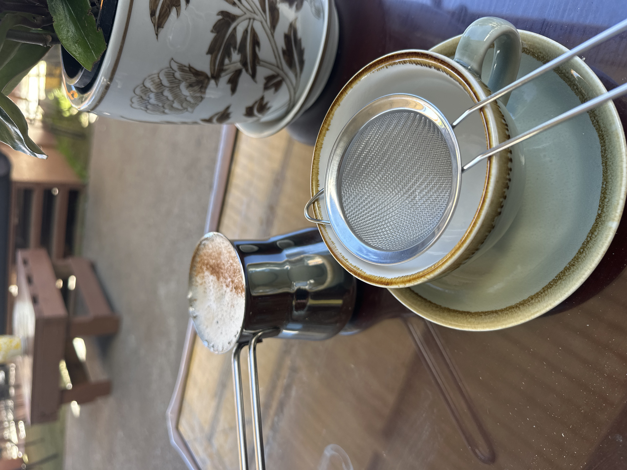 The image depicts a close-up view of a coffee setup on a dark, reflective table, likely outdoors. In the foreground, a light blue teacup and saucer are positioned, with a silver mesh strainer resting on top of the cup. To the left, a small, metallic coffee pot, known as a cezve or ibrik, contains a frothy coffee beverage, dusted with cinnamon or cocoa powder. In the background, out of focus, a wooden table and chairs are visible, suggesting a cafe or outdoor seating area. A potted plant in a decorative ceramic pot is situated to the right of the coffee pot. The lighting suggests it is daytime, with sunlight filtering through. The scene is likely set in Bellingen, Australia, given the context.