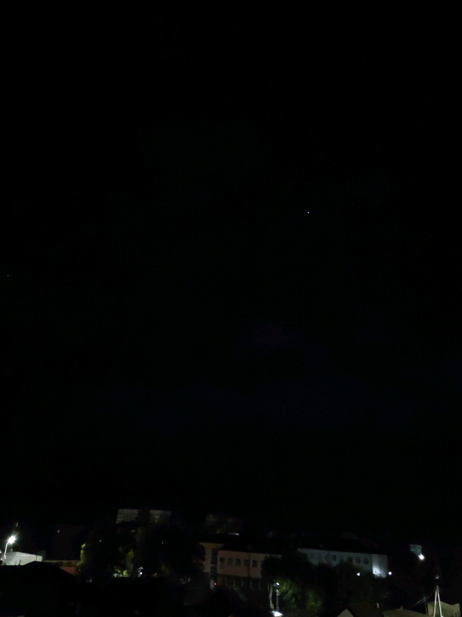 The image is a night time cityscape with a dark sky. In the upper portion of the sky, two small, bright points of light are visible, likely stars or planets. The lower portion of the image shows the silhouettes of buildings and trees, with some lights from the buildings and streetlights illuminating the scene. The city appears to be Mahilyow, Belarus. The scene is a typical urban night view.