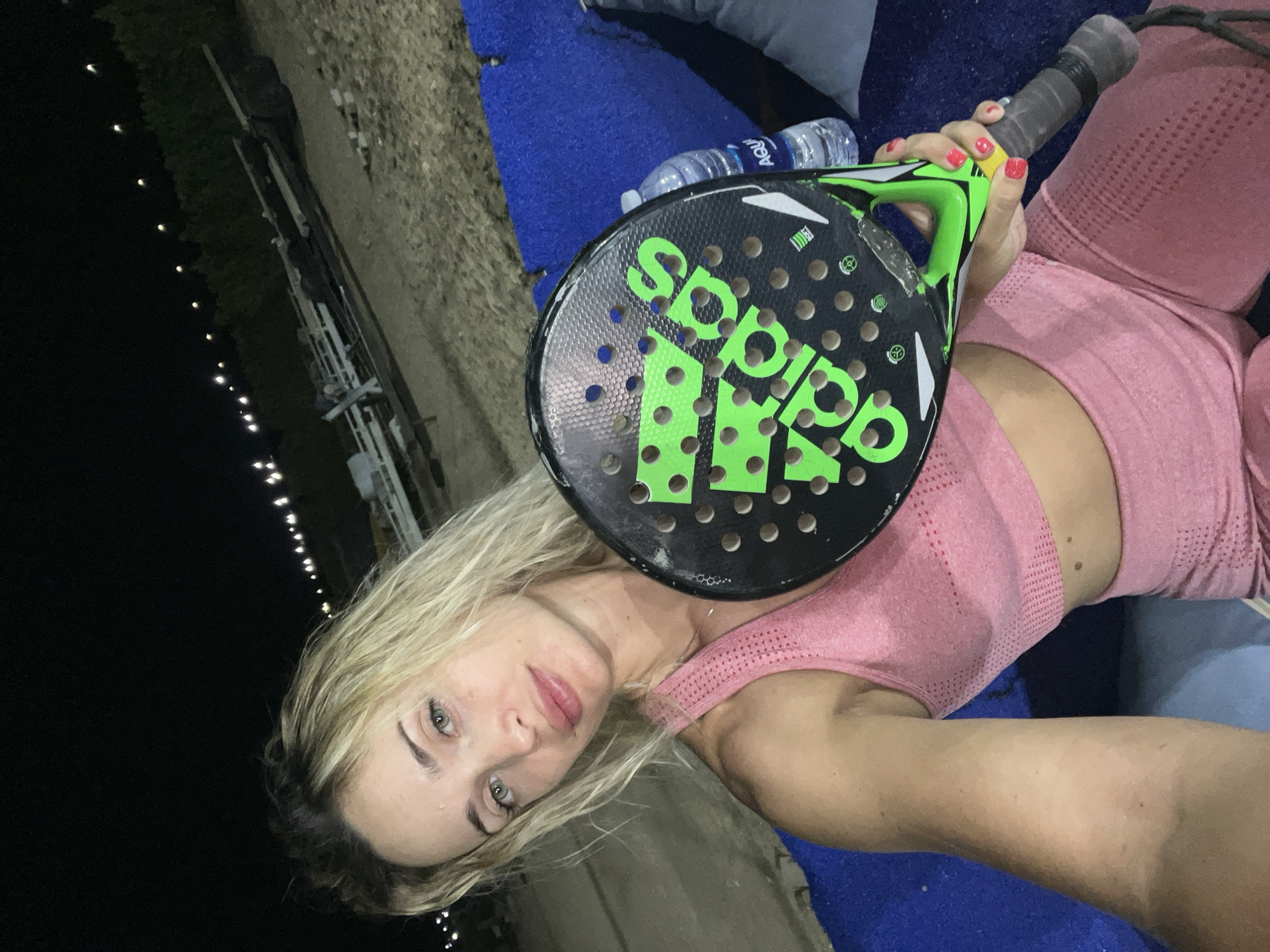 The image features a woman holding a black and green Adidas padel racket. She is wearing a pink athletic top and appears to be at an outdoor sports facility at night. The background is dark, with a line of lights visible in the distance, suggesting the facility might be illuminated for evening play. A blue mat and a bottle of water are also visible. The woman is looking towards the camera with a neutral expression. The presence of the padel racket and athletic wear indicates a sporting context. The location is identified as Sharm El Sheikh, Egypt.