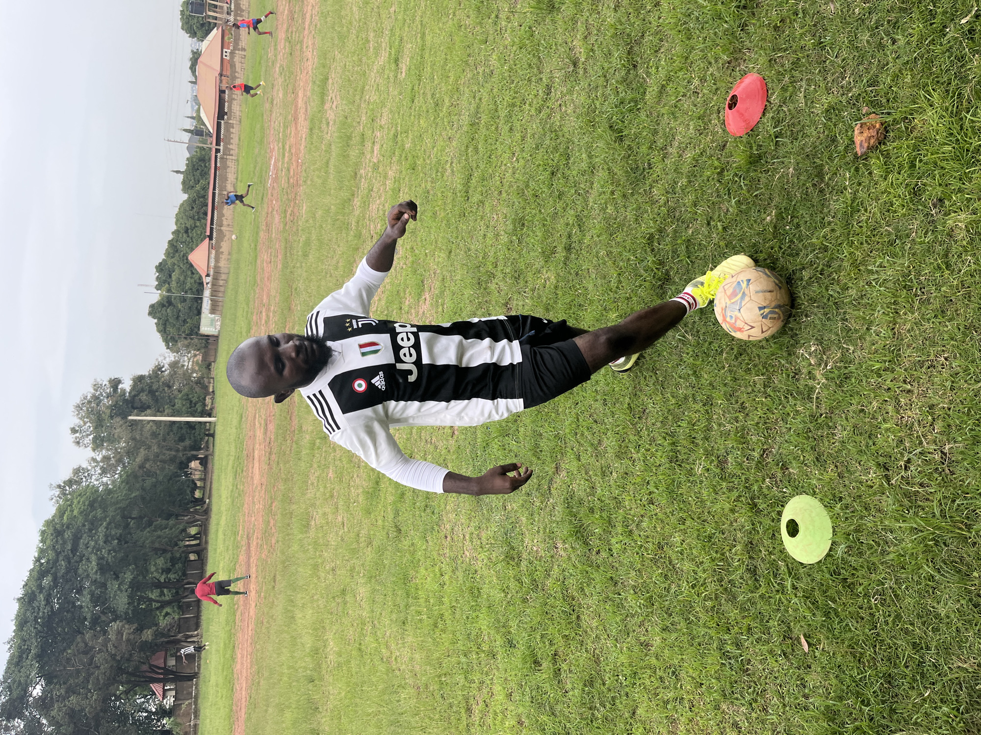 This media file depicts a group of people on a grassy field, likely a football pitch, in Jos, Nigeria. The primary subject is a man in the foreground, dressed in a black and white striped jersey with the "Jeep" logo and Adidas branding visible. He is in motion, with his right leg extended towards a football. Yellow cleats are visible on his feet. Other individuals are scattered across the field, some appearing to be engaged in football activities. In the background, there are buildings and trees, suggesting a semi-urban or recreational setting. Two conical markers, one red and one green, are placed on the grass, commonly used in sports training. The ground appears to be a mix of green grass and reddish-brown dirt. The lighting suggests it is daytime, and the weather appears overcast, contributing to a slightly muted color palette. No overt emotions are discernible from the individuals. The scene captures a moment of athletic practice or play in an outdoor environment.