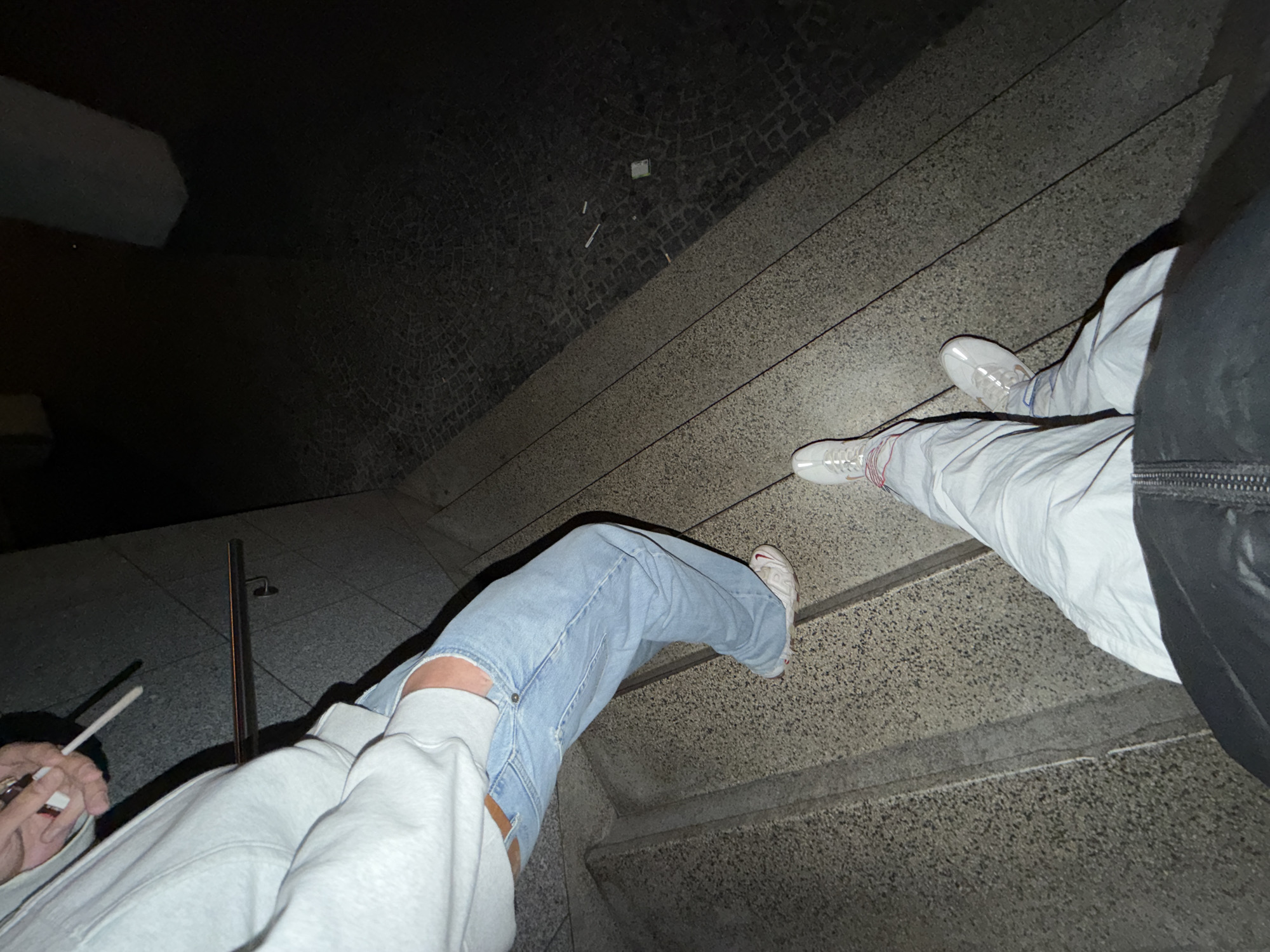 The image presents a low-light, downward-angled view of a concrete staircase in Vila Real, Portugal, within what appears to be an urban pedestrian passage or underpass. In the midground, a person is positioned on the stairs, wearing a light grey hooded sweatshirt, light blue distressed jeans, and white sneakers with a distinctive sole. This individual's right hand holds a white cigarette and a small, dark rectangular object, possibly a lighter or cigarette pack, near their hip. A dark, tubular handrail is visible against the textured wall to the right of this person. In the foreground, the legs and feet of a second individual are visible, wearing light-colored trousers and white sneakers with prominent red lacing and accents, indicating they are positioned higher on the staircase. The ground level to the lower right consists of dark, textured paving, possibly cobblestone, on which several discarded cigarette butts and a small, green rectangular object are visible.