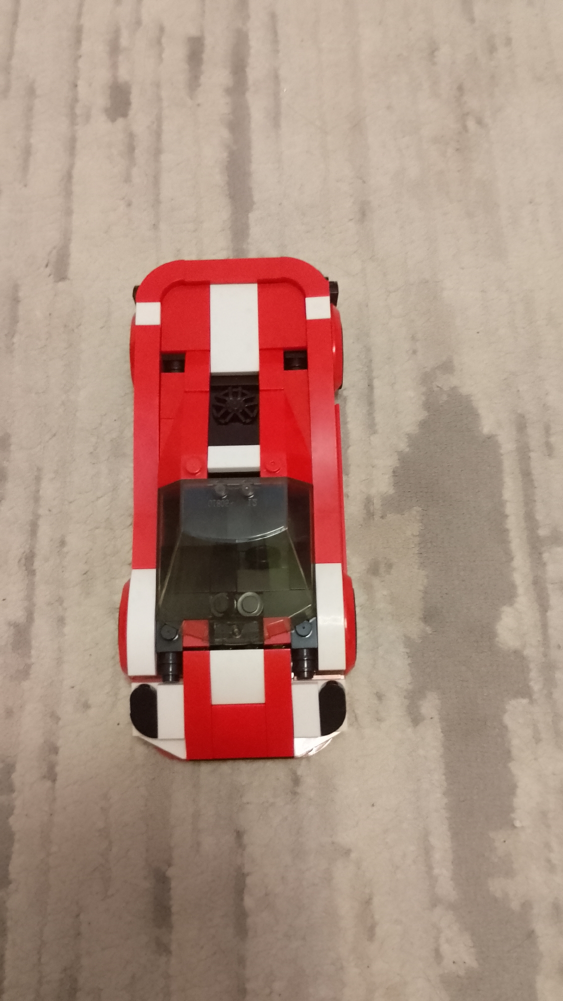 An overhead shot displays a red and white toy car, likely constructed from building blocks such as LEGO, resting on a light-colored, patterned carpet. The car is the primary subject, oriented vertically in the frame with its front facing downwards. It features a predominantly red body with a distinctive white stripe running down its center from front to back, and additional white elements on its sides and rear bumper.
The car's design suggests a sports or race car, characterized by its sleek, elongated form. It has a dark, translucent canopy or windshield over the cockpit area, under which some internal block structures are visible. On this canopy, faint, small text or numbers can be discerned, appearing to read "D280P" and possibly "T3" or "73". Black components, including what appear to be wheels or wheel wells, are visible at the sides and rear.
The setting is an indoor space, indicated by the carpeted floor. The carpet features a subtle, irregular striped or mottled pattern in muted tones of beige and light grey. No activity is taking place; the car is stationary on the carpet. The lighting suggests an indoor environment but does not provide cues about the time of day.