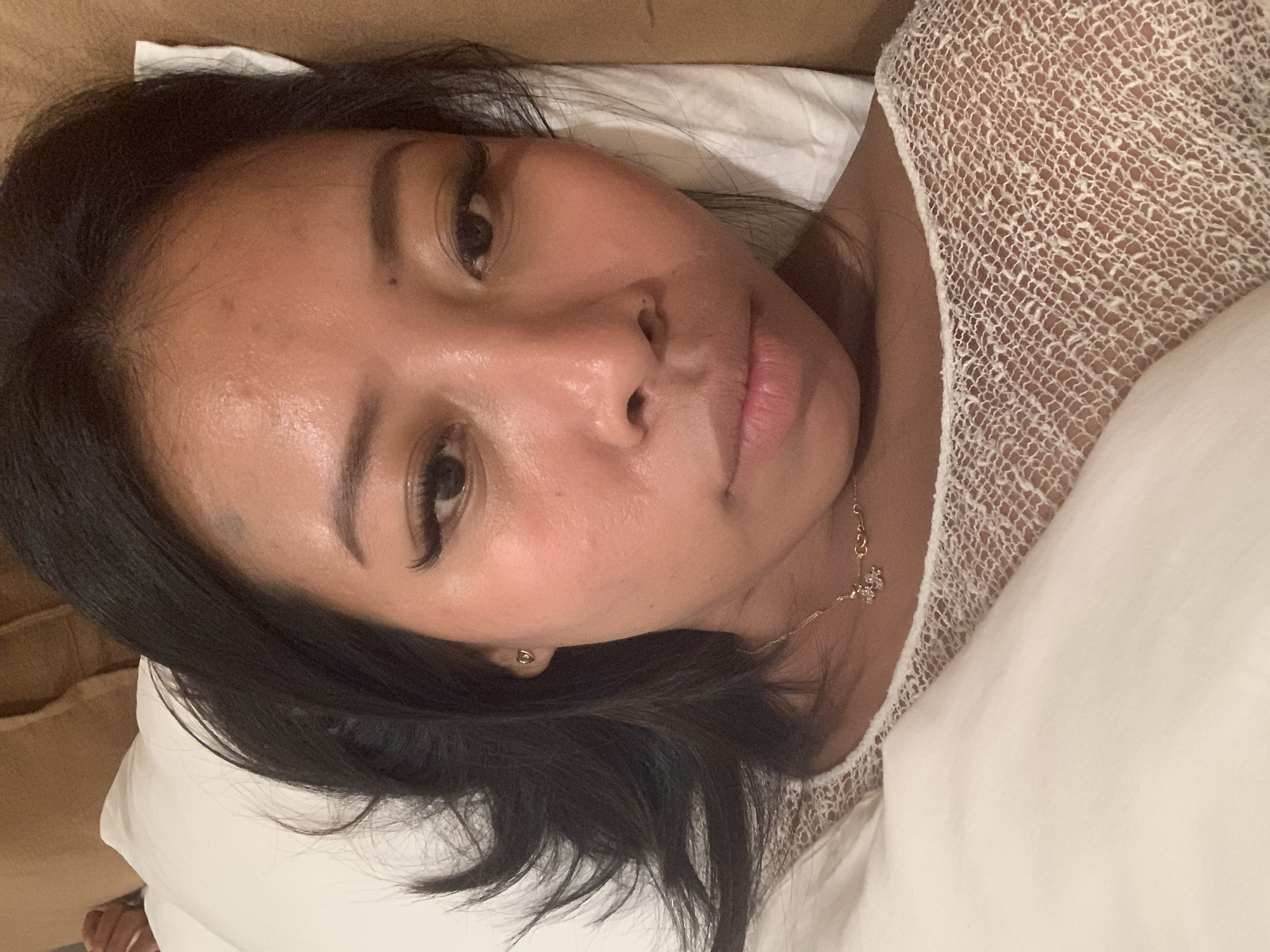 The media file shows a close-up, rotated, over-the-shoulder shot of a person lying down in what appears to be a bed. The person has dark hair and is wearing a white, intricately patterned top. A gold necklace with a small pendant is visible on their neck. The background consists of pillows and bedding, with a hint of a brown surface, possibly a headboard or furniture. The lighting suggests it could be nighttime or indoors with soft lighting, and there are no indications of weather conditions. The overall mood is relaxed and intimate, with the person's face visible and a neutral expression. No text is present in the image. The scene is likely set in a bedroom in North Kuta, Indonesia.