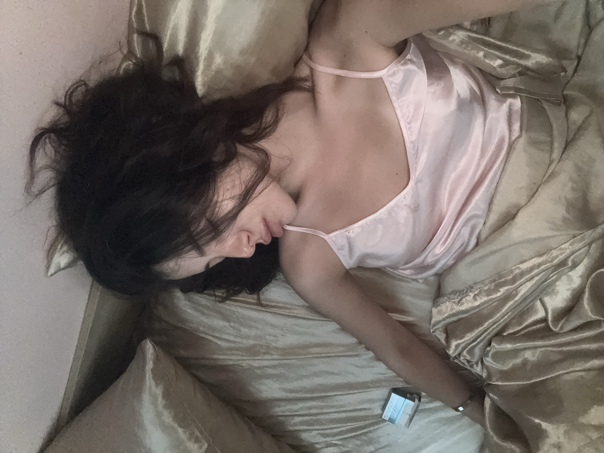 A woman is lying in bed, asleep. She has dark hair and is wearing a light pink camisole. The bed is covered in satin sheets and pillows. A small electronic device is on the bed next to her left arm. The scene appears to be a bedroom. There is no information to indicate the location in the city of Plovdiv, Bulgaria.