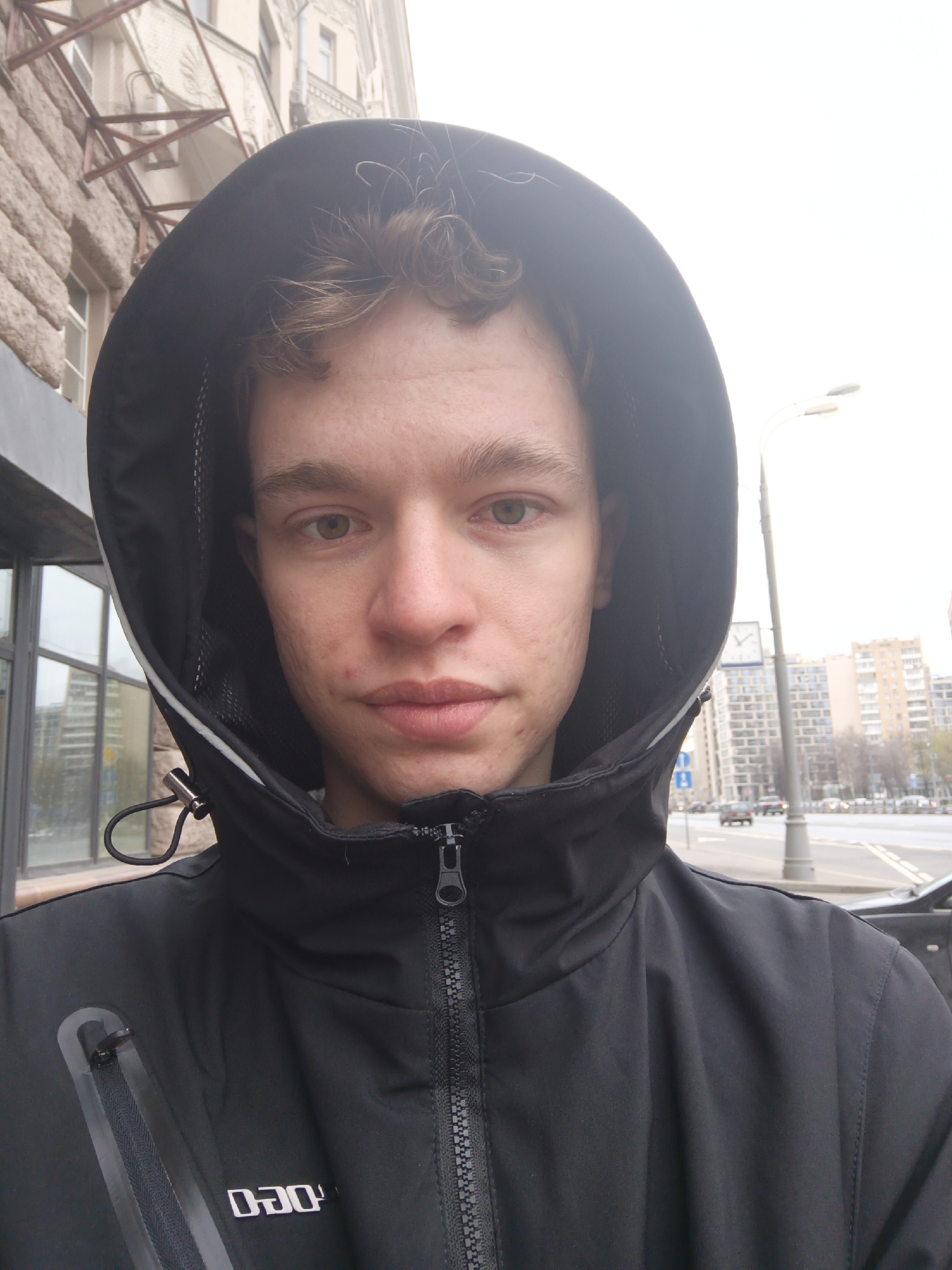 A young Caucasian male is centered in a close-up shot, wearing a black hooded jacket zipped up to his neck. His hair is light brown and curly, and he has blemishes on his face.
The background shows a city street. Buildings are visible across the street, one appearing to be made of light-colored bricks. A street light is also visible. Cars are driving on the street in the distance. The time is probably daytime, and it looks like it could be Moscow, Russia.