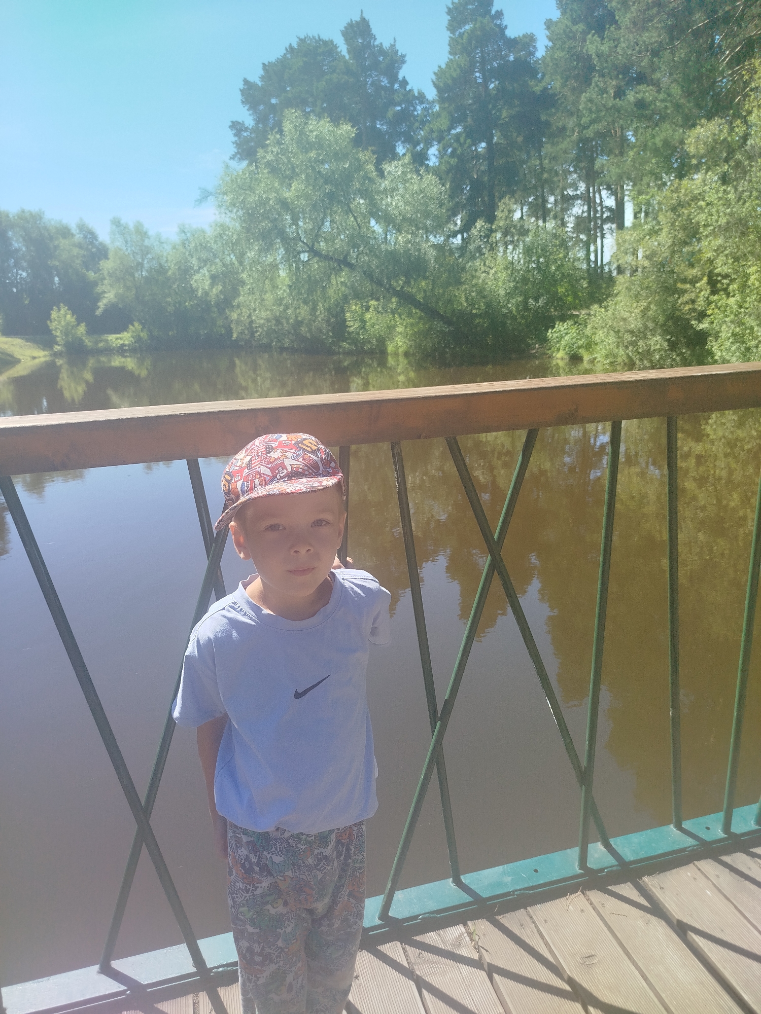 A young boy wearing a light blue t-shirt and patterned shorts stands on a wooden bridge, looking at the camera. He is wearing a colorful baseball cap. The bridge has a wooden railing and green metal supports. Behind him is a body of water, likely a river or lake, with reflections of trees on its surface. The background features lush green trees and a clear blue sky. The scene appears to be in an outdoor recreational area.