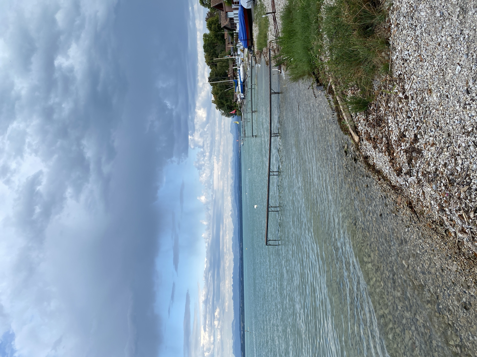 This image depicts a lakefront scene with a view of the Jura Mountains in the distance, under a cloudy sky. In the foreground, a pebble beach meets the clear, shallow water, with a wooden pier extending into the lake. Several boats, some covered with tarps, are moored along the shore, and a few buildings with red roofs are visible amongst trees. A person on a paddleboard is present on the water. The overall impression is of a calm, natural setting in a lakeside town.