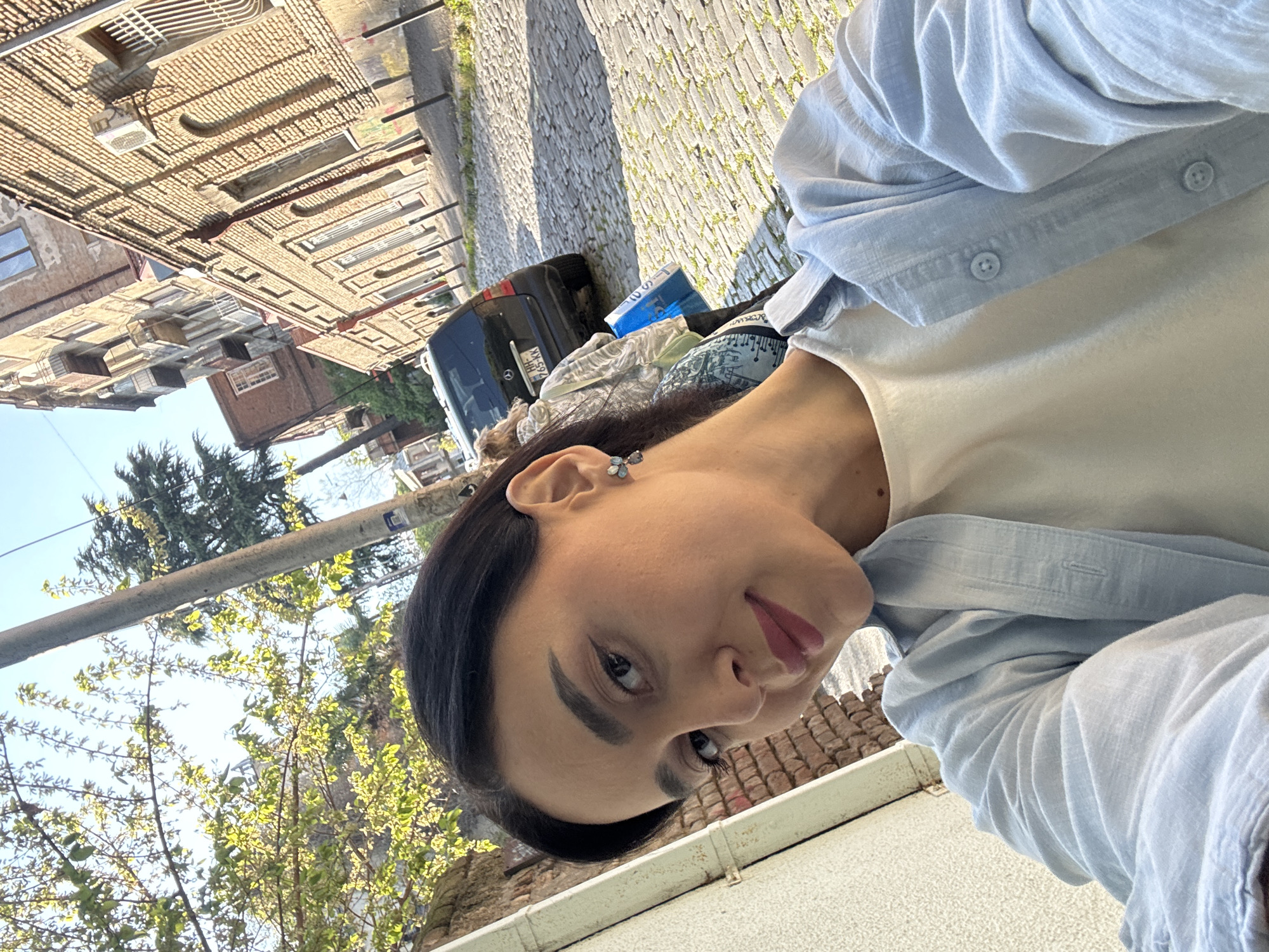 A young adult woman poses for a selfie outdoors, viewed from a slightly low angle. She has dark hair, dark eyebrows, and pink lipstick, and wears a light blue button-up shirt unbuttoned over a cream or off-white top. A small, light blue or silver earring is visible in her left ear. Her expression is calm, with a slight smile.
The setting is an urban street in Tbilisi, Georgia, under a clear blue sky. In the background, there are multi-story buildings with brick or stone facades and visible window units or balconies, suggesting older European architecture. A dark-colored car, possibly an SUV, is parked further down the street, partially obscured. Its license plate, "XX 695 KK," is visible. The ground is made of cobblestones. Lush green trees with new leaves indicate spring or early summer, and bright sunlight suggests it is daytime. Some bags or items are visible near the parked car. A utility pole or horizontal bar crosses the upper left part of the background.