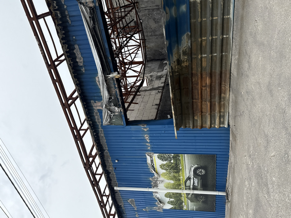 The image shows a partially destroyed building in Horenka, Ukraine (50.56, 30.309). The building is made of blue corrugated metal and has a rusty metal roof. The roof has been partially destroyed, and the building appears to have been damaged by fire or an explosion. The image shows a black car poster on the building, the poster features a black car in the foreground, and trees in the background, with the word "BANDIT" on the side of the car. The building is located in a residential area, and there are no people present. The building is surrounded by rubble and debris. The image shows the destruction caused by the Russian invasion of Ukraine.