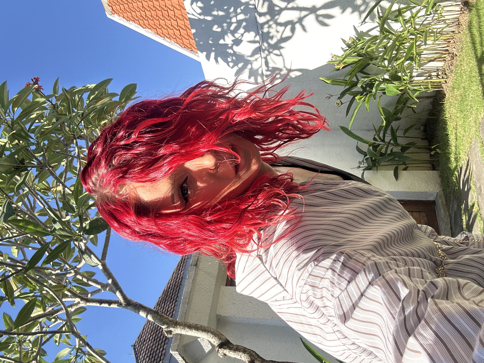 The image features a person with vibrant red, curly hair, prominently angled across the frame. Their face is partially visible, with one eye catching the sunlight and a subtle, friendly expression. They are wearing a light-colored shirt or dress with thin stripes, and a gold-colored chain is visible on their shoulder.
The scene is outdoors on a bright, sunny day. A clear blue sky is visible in the upper-left portion of the image. Lush green foliage, including a tree with thick branches and broad leaves, possibly a frangipani given the faint red blossoms, occupies the lower-left and central areas. Part of a building with an orange-tiled roof is visible in the top-left corner.
On the right side of the frame, a white wall is adorned with distinct, intricate leaf shadows cast by the intense sunlight, indicating strong illumination. Green plants line the base of this wall, alongside a patch of well-maintained green grass in the bottom-right. The bright conditions, clear sky, and tropical vegetation suggest a warm climate, consistent with the North Kuta, Indonesia context. The time of day appears to be mid-morning or afternoon, judging by the sharp shadows. No visible text is present.
