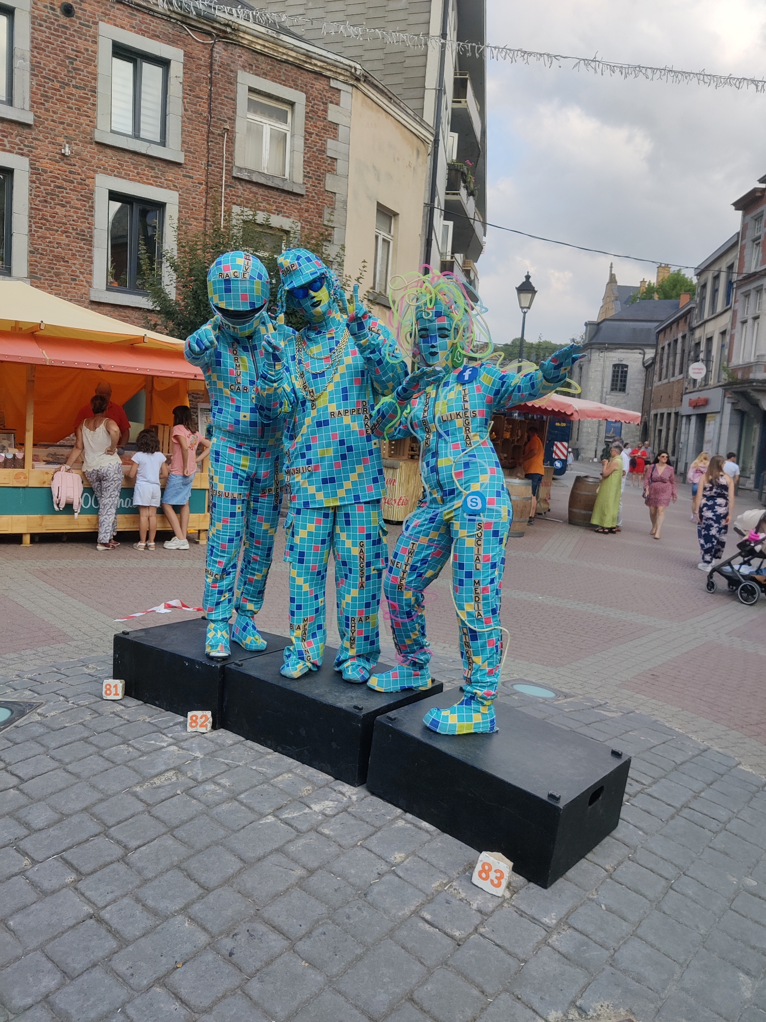 The image depicts a street scene in Marche-en-Famenne, Belgium. In the foreground, three performers dressed in colorful, checkered costumes stand on black platforms. The costumes are designed to resemble mosaic patterns and feature text related to social media platforms and internet terms such as "FACEBOOK," "TWITTER," "LINKEDIN," "INSTAGRAM," "MUSIC," "GANGSTA," and "RAPPER." One performer on the far left wears a helmet resembling a racing helmet, with the word "RACE" visible. The performer in the center wears sunglasses and makes a peace sign with their fingers. The performer on the right has colorful curly hair-like attachments and is gesturing with an outstretched arm. In front of the platforms, small numbered blocks are placed on the cobblestone pavement, with "81" to the left of the first platform, "82" between the first and second, and "83" to the right of the second.
In the background, there are buildings with brick facades and windows, typical of a European street. A market stall with an orange awning is visible to the left, with several people interacting with it. Further down the street, other pedestrians are walking, and a baby stroller is visible. The sky is overcast with a few scattered clouds. The overall scene suggests an outdoor event or street performance.