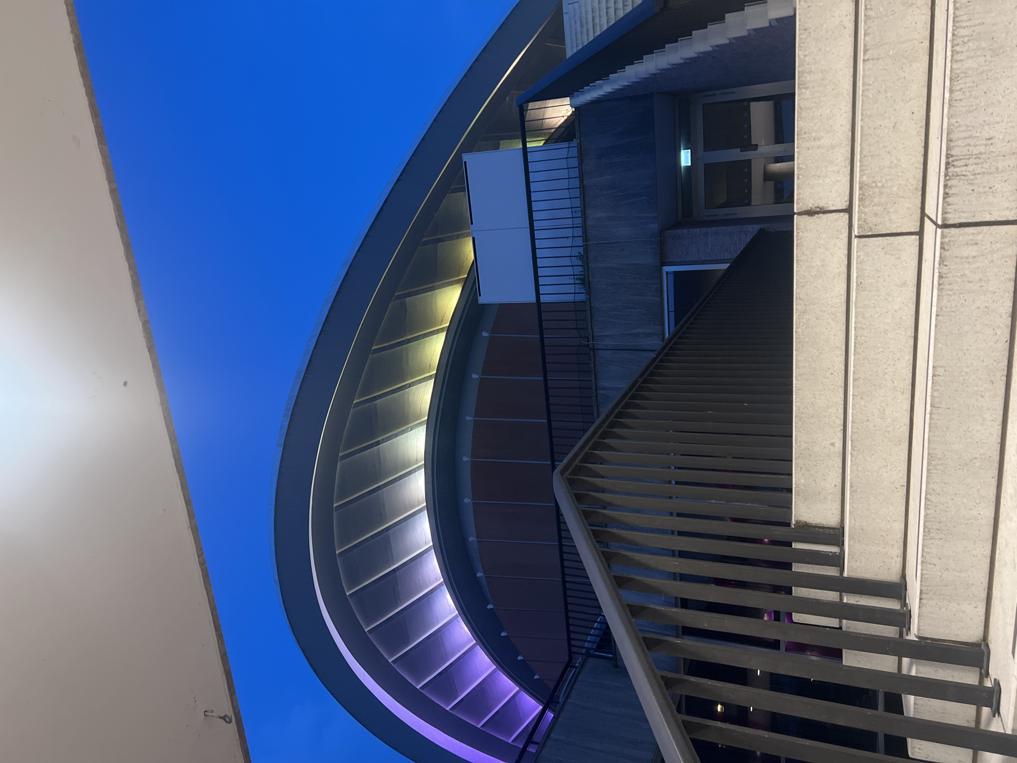 The image captures the illuminated exterior of the Philharmonie in Berlin, Germany, at dusk. A grand, curved roof, outlined by purple and yellow lights, dominates the upper portion of the frame. Below the roof, a darker section of the building features tinted windows and what appear to be wooden panels. A black metal railing runs along a concrete staircase that leads upwards. The stairs themselves are made of concrete and show some signs of wear. The sky above is a deep, clear blue, transitioning to a darker hue as night approaches. No people are visible in the scene.