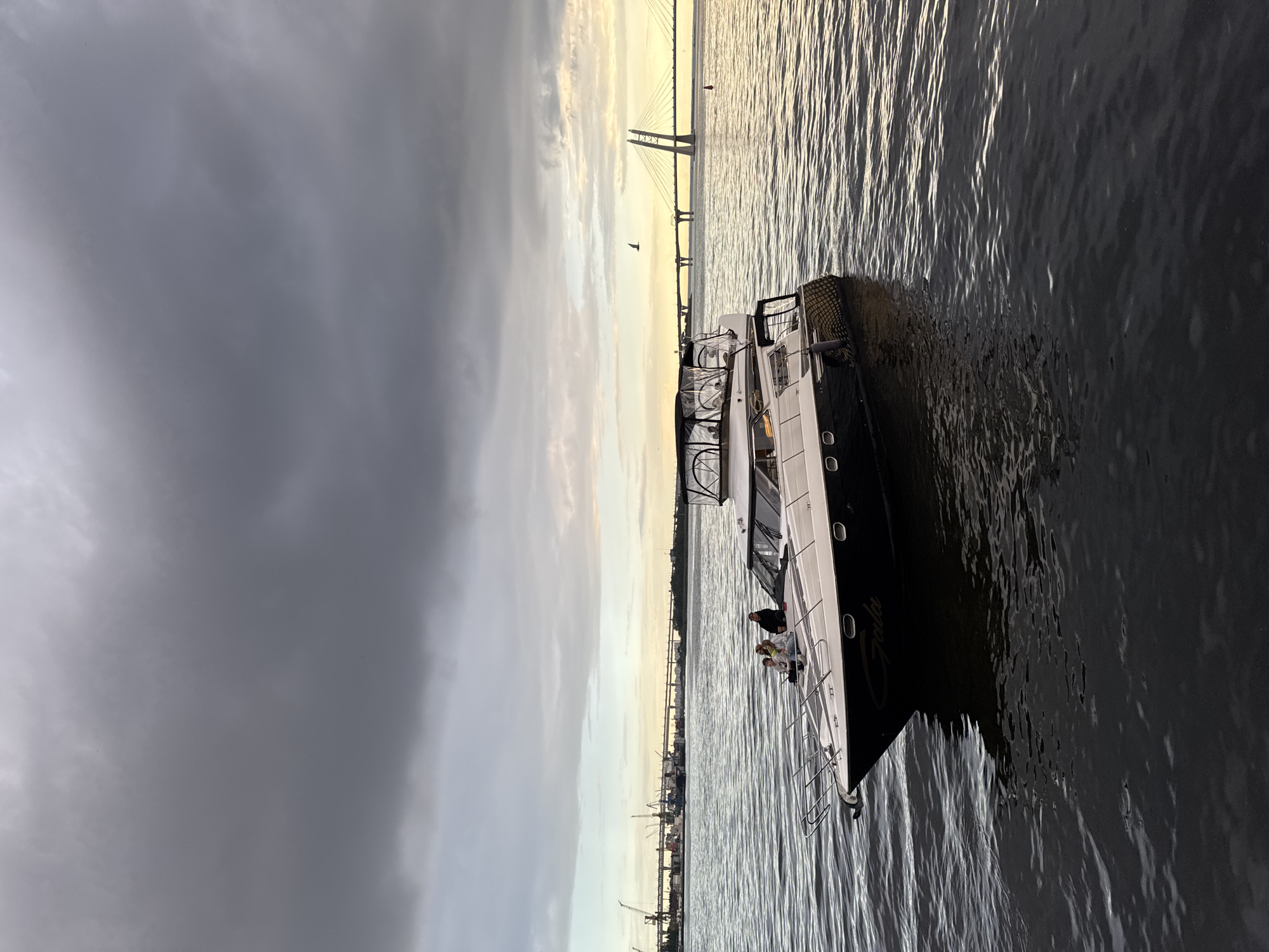 The image captures a motor yacht named "Gala" docked on a body of water, likely the Neva River in Saint Petersburg, Russia, under an overcast sky. The yacht is white with black accents and features a canopy structure. Three individuals are seated on the bow of the yacht, facing left. In the background, a large cable-stayed bridge, identified as the Western High-Speed Diameter, spans the water, with industrial cranes and buildings visible further in the distance on the left. The water is choppy, and the light suggests it is either early morning or late evening.