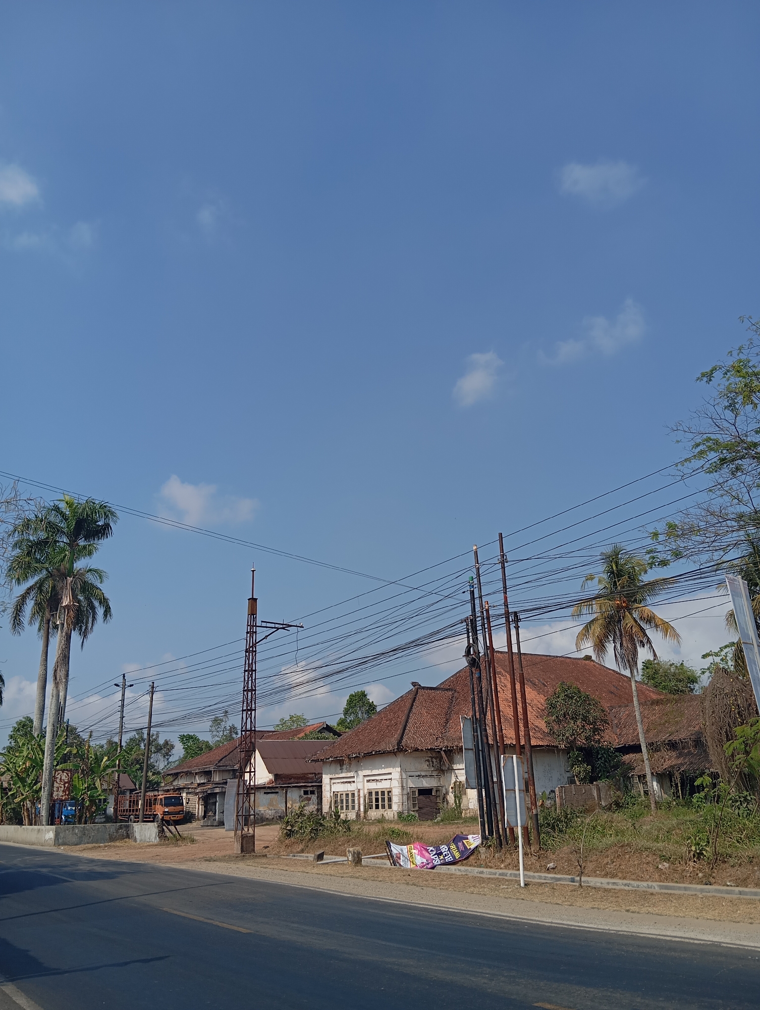 The image depicts a roadside scene in Sokaraja, Indonesia, under a clear blue sky with scattered white clouds. On the left, two tall palm trees stand next to a street. Across the street, a cluster of old buildings with red tile roofs are visible. One building appears to be a long, single-story structure with multiple windows and a somewhat dilapidated appearance. In front of these buildings, there are several utility poles with numerous overhead wires. An orange truck is parked in front of one of the buildings, and a large banner lies on the ground in front of another. The ground in front of the buildings is covered with dry grass and vegetation. The road in the foreground is paved and appears to be a two-lane road. The overall impression is of a rural or semi-rural setting in a tropical climate.