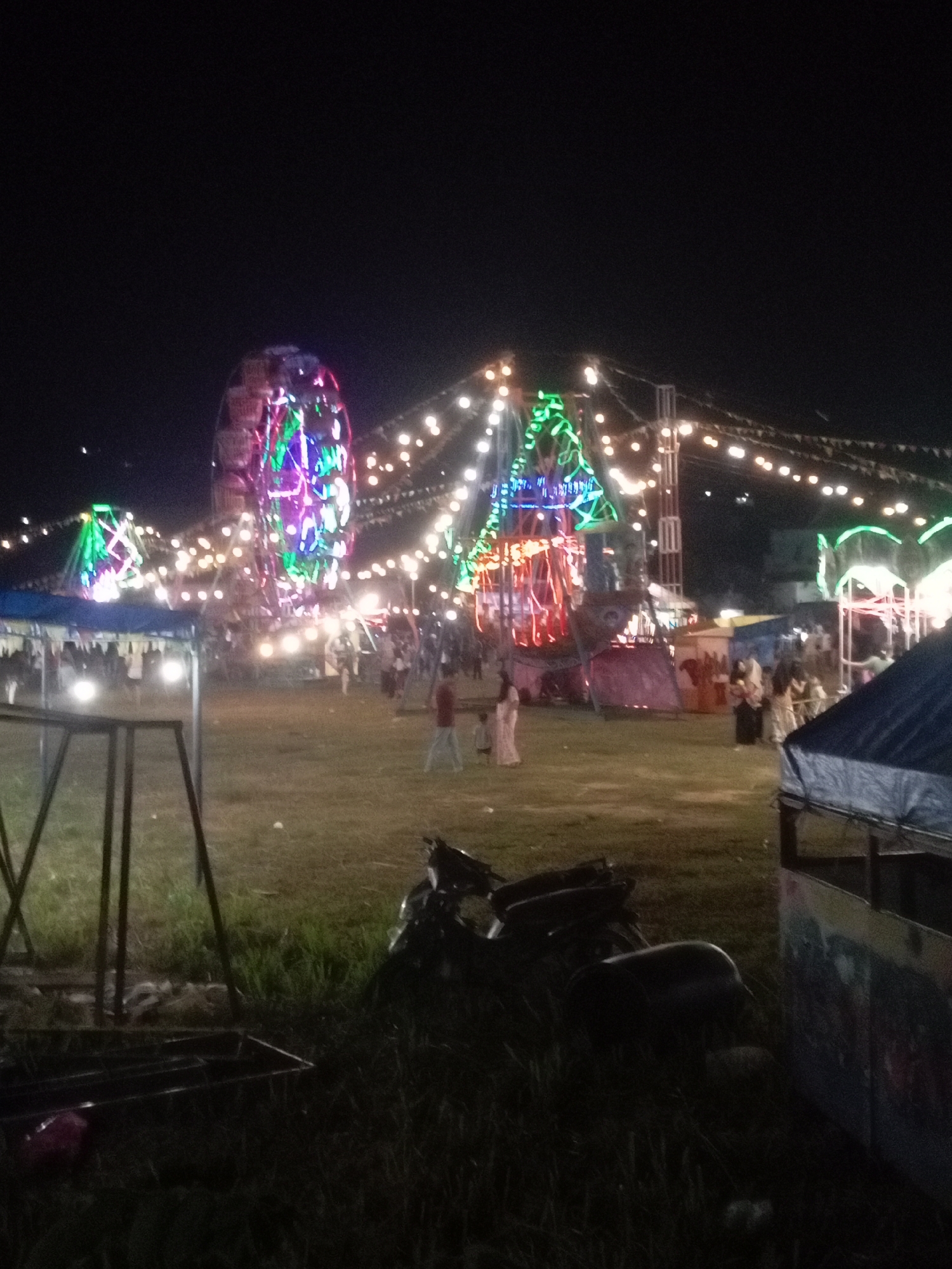 The image depicts a night scene at a fairground, likely in Batam, Indonesia, given the context of the request. Prominently featured is a brightly lit Ferris wheel, with colorful lights illuminating its structure. Several other amusement rides, also adorned with festive lights, are visible in the background, contributing to a carnival atmosphere. A string of small, circular lights spans across the fairground, adding to the ambiance. The ground is covered in grass, and there are various temporary structures and tents. Several people are present, some walking and others gathered around the rides and stalls. In the foreground, a motorcycle is parked on the grass, and a dark metal frame, possibly part of a swing set or another ride, is also visible. The overall impression is that of a bustling night market or fair.