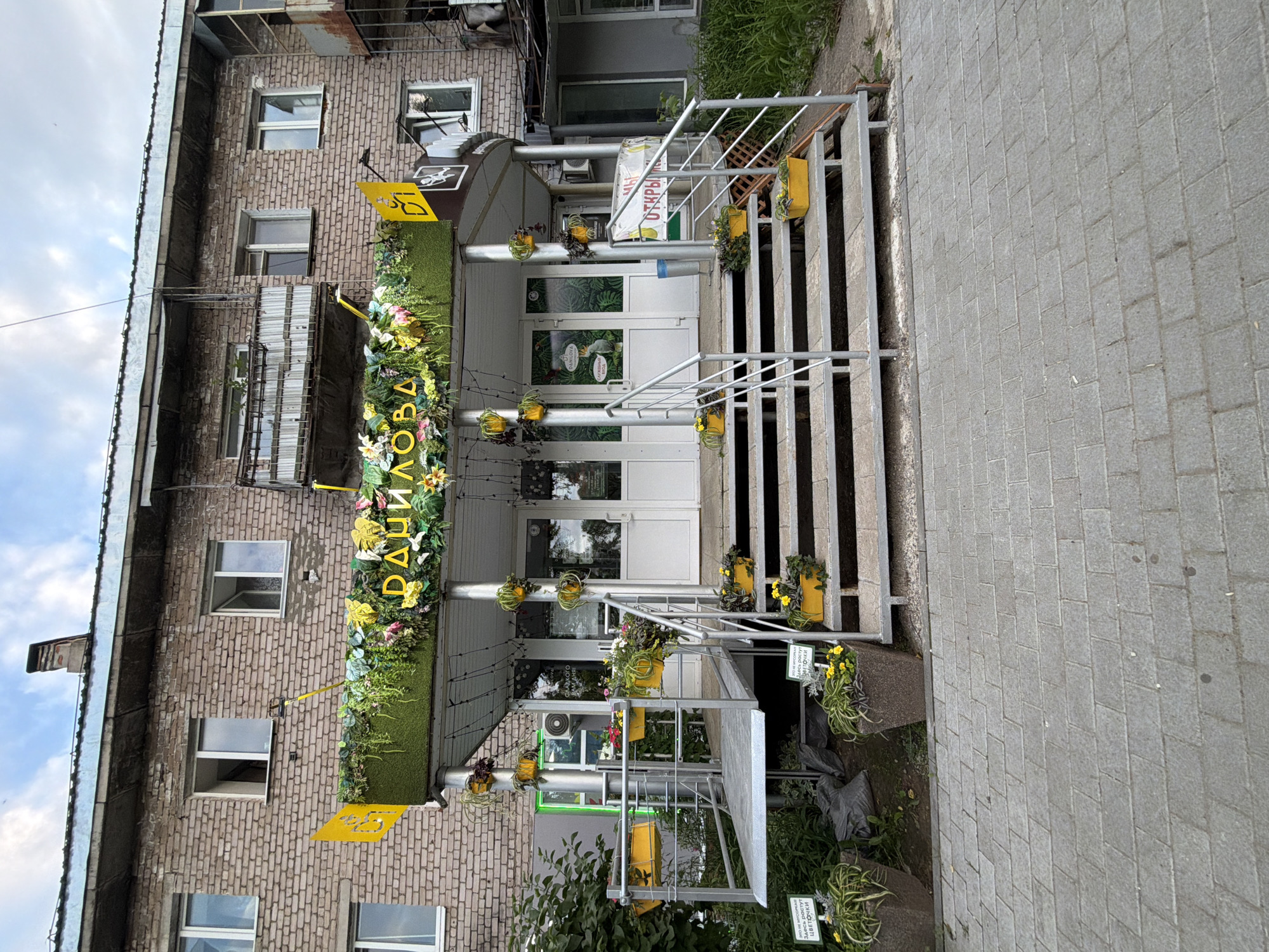 The image shows the entrance to a flower shop in Chaikovsky, Russia. The shop has a vibrant display of artificial flowers and greenery above its entrance, with the shop's name, "ДАНИЛОВА," written in large yellow letters. There are two yellow signs on either side of the entrance, one featuring a stylized logo with Cyrillic text. White doors lead into the shop, and one of the doors has a depiction of a parrot. A ramp and a set of metal stairs with handrails lead up to the entrance. Yellow planters filled with yellow and pink flowers are placed along the stairs and on a platform near the ramp. The building is made of brick, and there is a balcony on the second floor. The ground in front of the entrance is paved with gray rectangular bricks, and there is some greenery and planters on either side of the walkway. A sign on one of the planters reads "ЭТО НЕ МУСОРКА. ЗДЕСЬ РАСТУТ ЦВЕТОЧКИ" which translates to "THIS IS NOT A TRASH CAN. FLOWERS GROW HERE." The sky is partly cloudy.