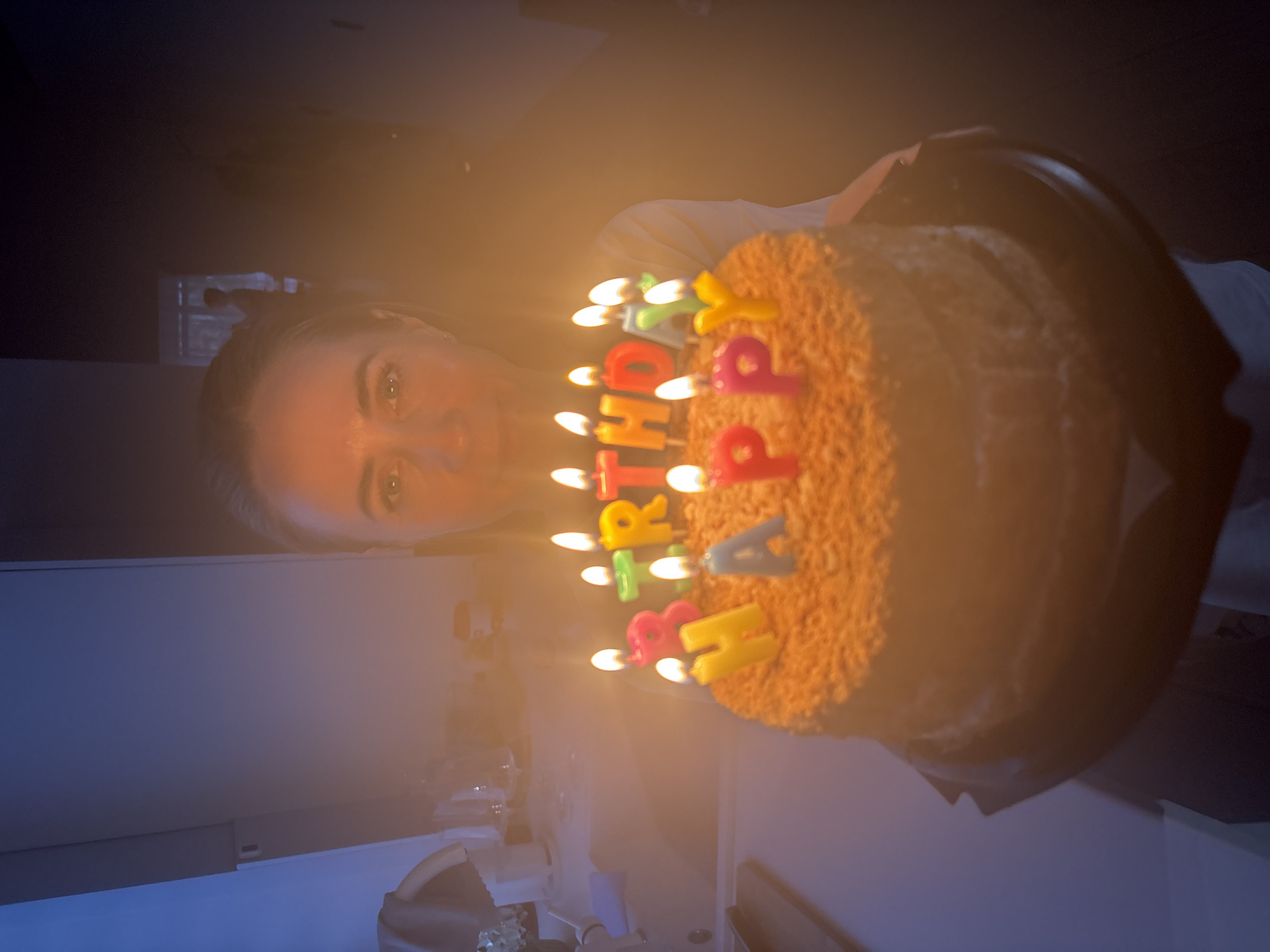 A woman is holding a birthday cake with lit candles that spell out "Happy Birthday". The cake is round and covered in what appears to be crumbled topping. The woman is looking at the camera. The location is the city of Chaikovsky, Russia. The background is dark, with a countertop and possibly kitchen appliances visible.