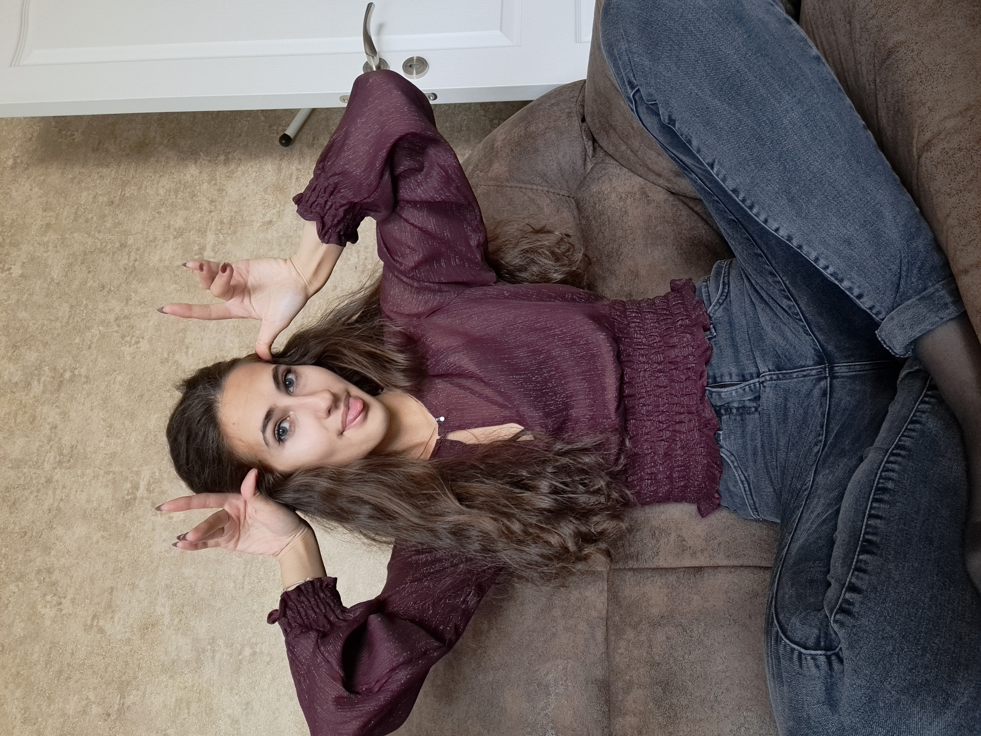 A young woman lies on a brown sofa. She wears a burgundy long-sleeved blouse and blue jeans. Her dark, wavy hair is loose. She is making a "peace sign" with both hands, placing them near her head as if they were horns. She is looking directly at the camera with a slight smile and her tongue poking out. A white door is visible in the upper left corner, suggesting an interior setting. There is no information to indicate that this photo was taken in Negovan, Bulgaria, or any specific location.