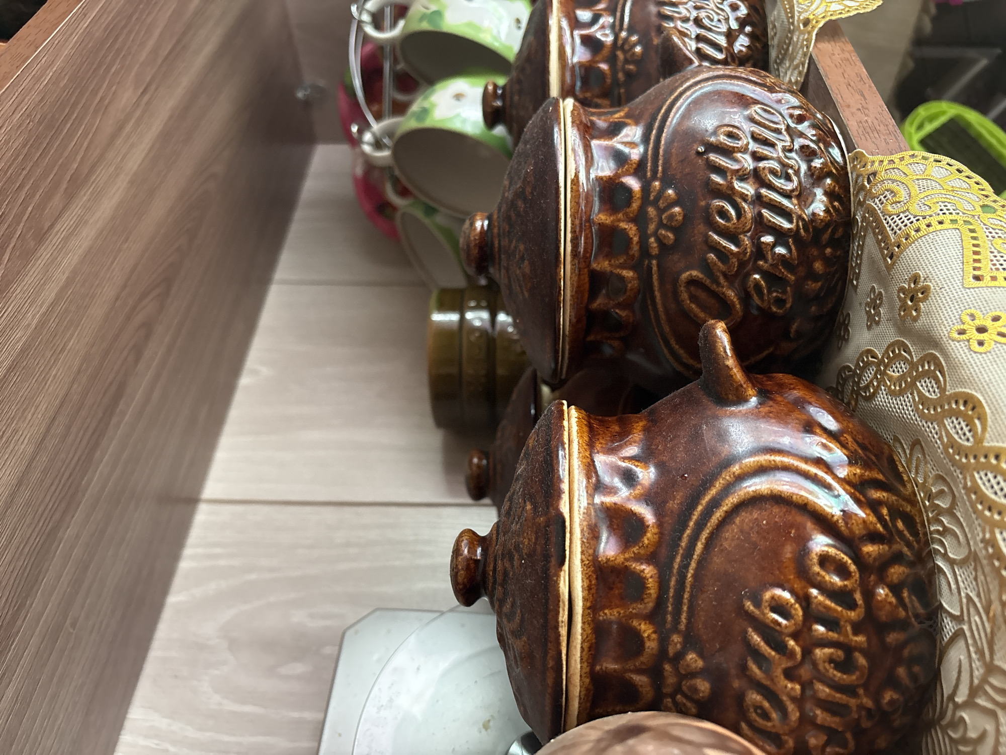 This image depicts a close-up view of several brown ceramic pots with lids, placed on a wooden shelf. The pots are decorated with embossed patterns and text. In the foreground, two pots are prominently displayed, one on the left and one on the right. The pot on the left has text that appears to read "Очень вкусно" (Ochen vkusno), meaning "Very tasty." The pot on the right also has similar text, partially obscured. Behind these pots, other items are visible, including what appears to be a white plate or bowl, a stack of green ceramic containers, and a rack holding green and white floral patterned mugs. A decorative embroidered cloth with yellow accents is laid out on the shelf beneath the pots. The overall environment suggests a kitchen or pantry setting. The image does not contain any people. The specific location in the city of Ветлужский, Russia, is not determinable from the image itself.