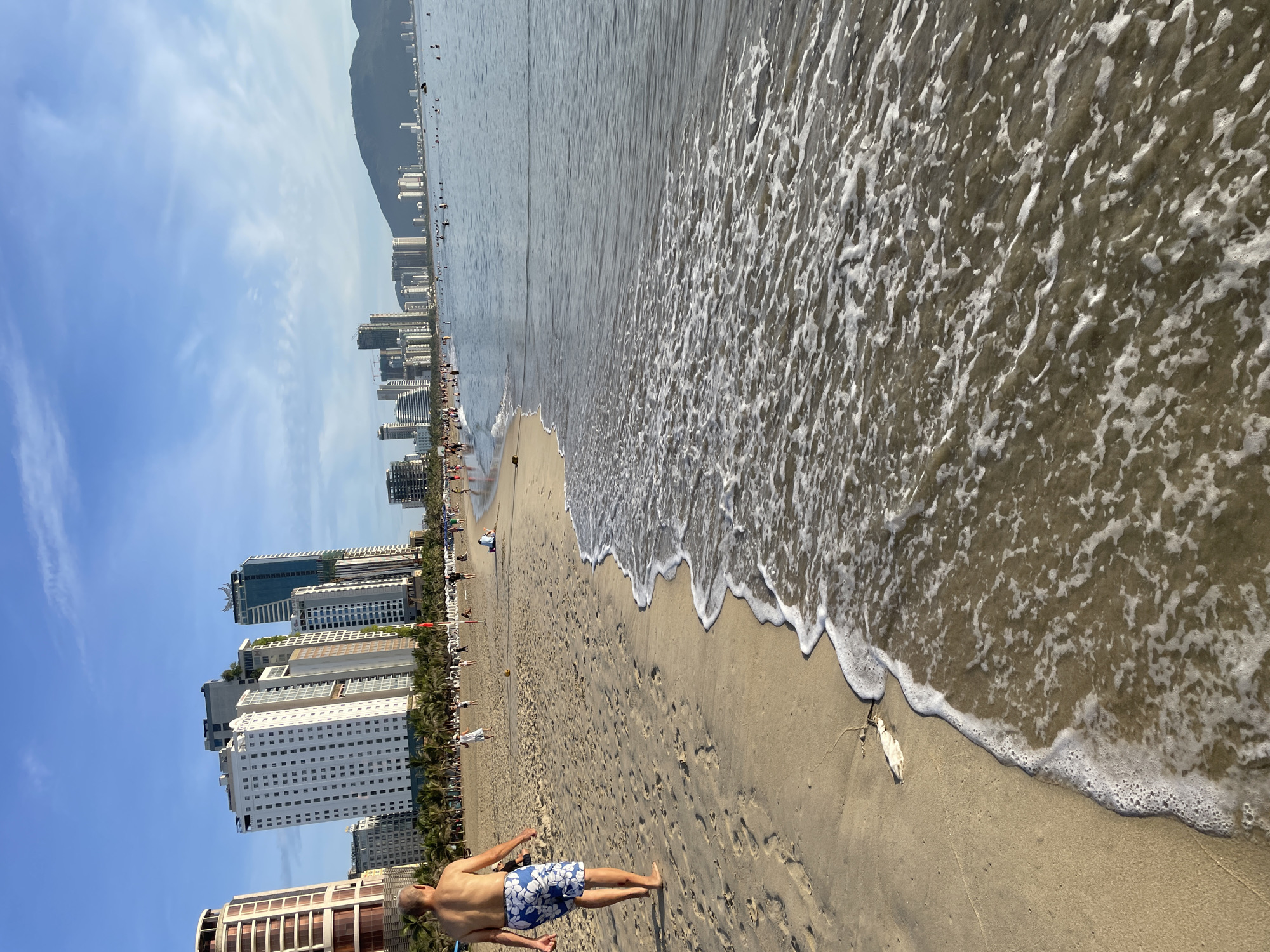 The image depicts a sunny day at My Khe Beach in Da Nang, Vietnam. In the foreground, a man with a bald head and floral swim trunks walks away from the camera on the sandy shore. The ocean waves gently lap at the beach, creating white foam. In the background, numerous high-rise buildings line the coast, characteristic of the Da Nang skyline. Several people are visible on the beach and in the water, enjoying the day. The sky is clear blue with a few wispy clouds. The scene captures a typical beach day with a blend of natural beauty and urban development.