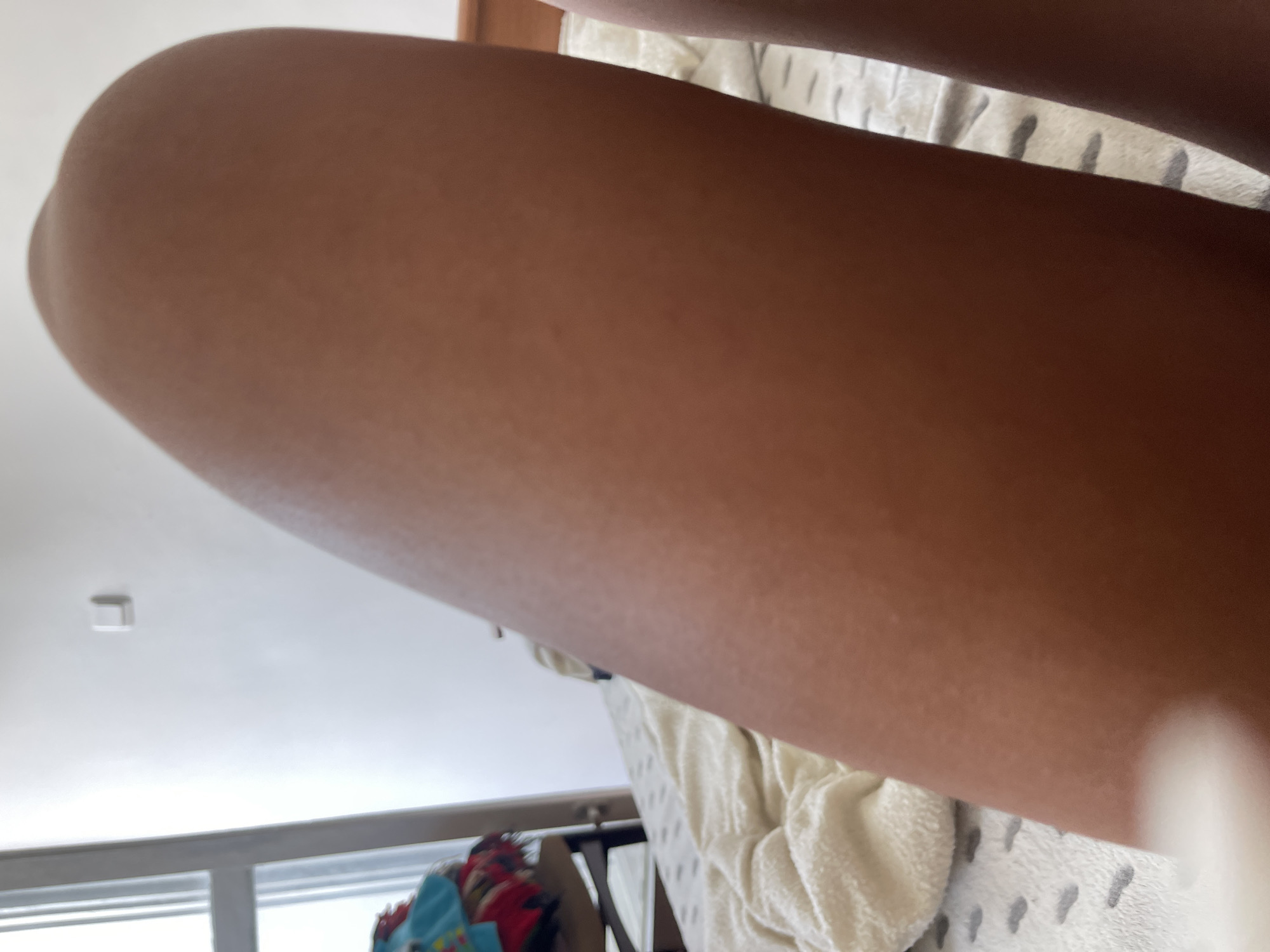 The image shows a close-up of a person's leg and thigh, likely in a bedroom setting. The skin is a medium brown tone with visible pores and a slight sheen. The background reveals a white wall with a light switch, a window with vertical blinds, and a bed with a textured white comforter. Some folded clothes are visible on a piece of furniture near the window. The overall impression is an indoor, domestic scene. There is no specific information in the image to suggest a location in Varna, Bulgaria.