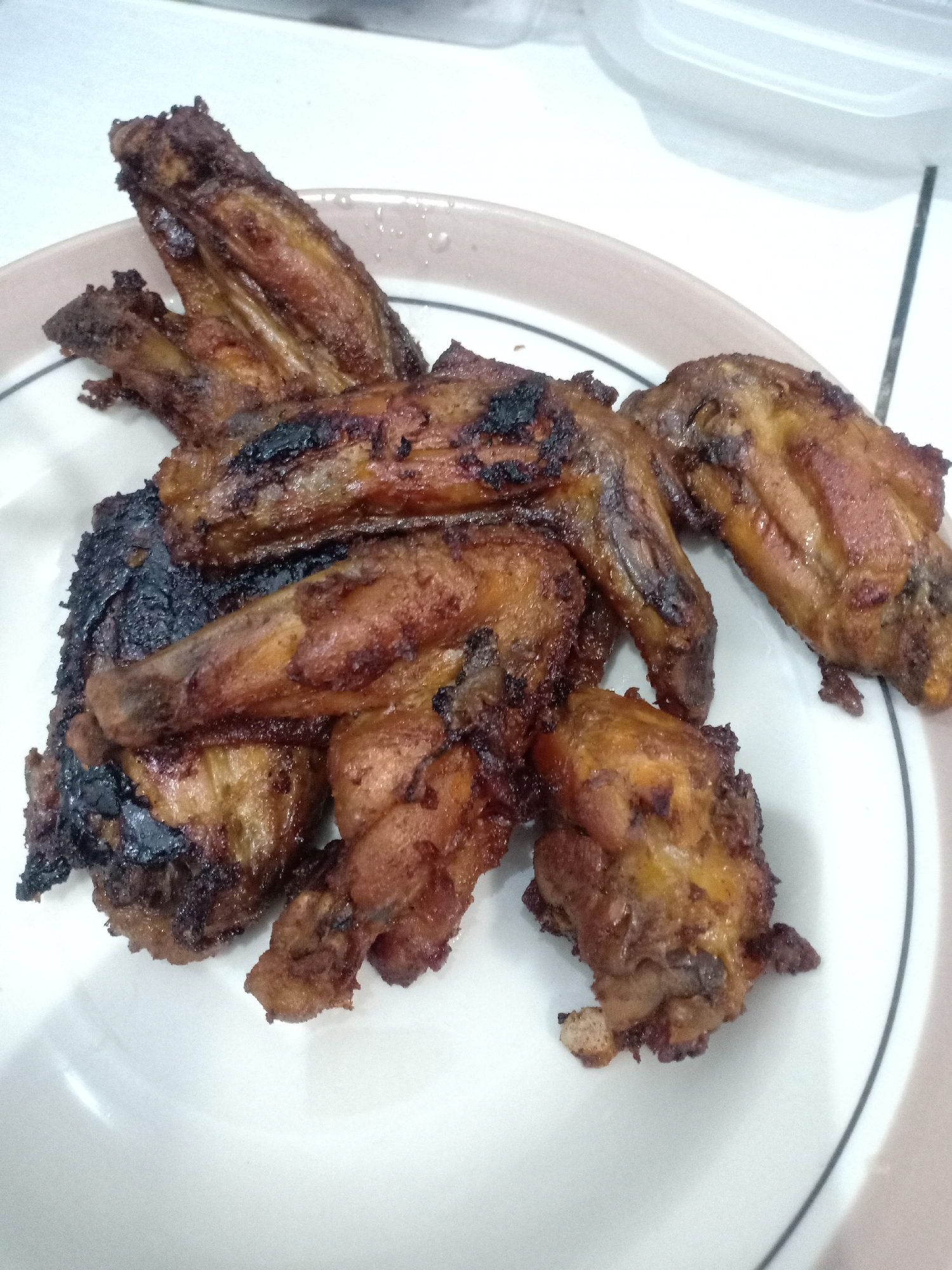 A close-up, overhead shot shows a pile of cooked chicken wings and smaller pieces of chicken on a white plate with a brown rim. The chicken is a deep brown color, with some areas appearing charred and crispy. The plate is on a tiled surface, with a white plastic container visible in the background. The lighting is artificial and somewhat harsh, casting shadows on the chicken. The image appears to be taken in a home kitchen setting in Semarang, Indonesia.