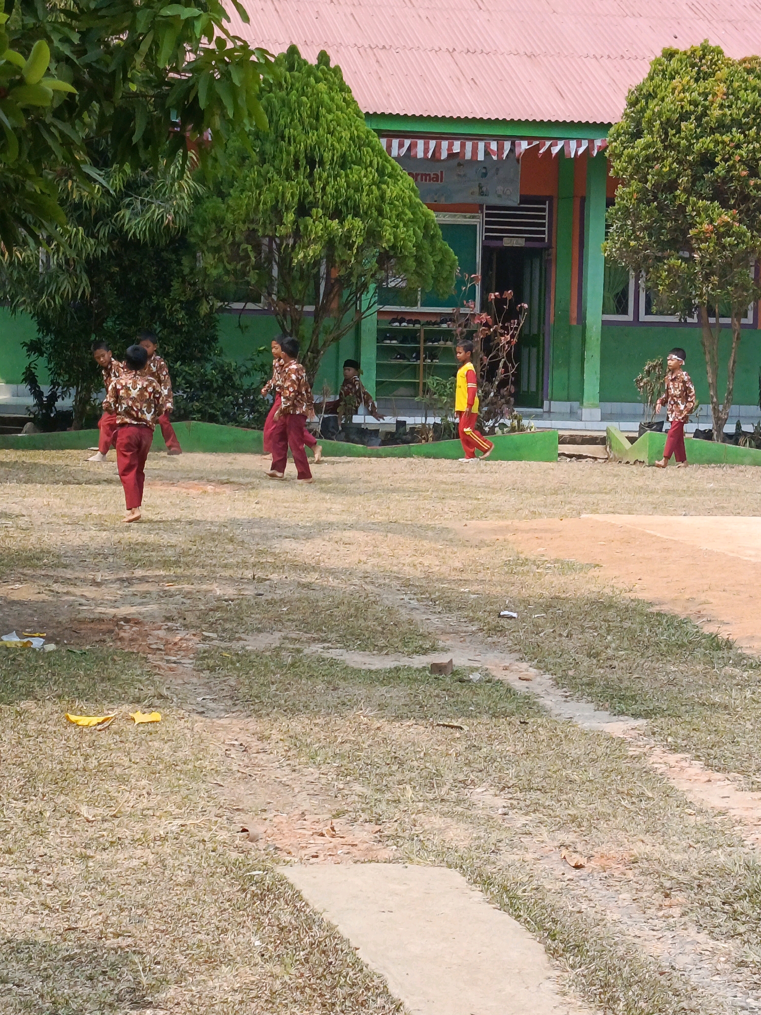 In Megang Sakti IV, Indonesia, a group of young children, likely students, are seen on a schoolyard during daylight. The children are dressed in similar uniforms: a patterned brown shirt with a cream-colored collar and a cream-colored pattern of what appears to be animals or floral motifs, paired with maroon pants.
There are at least six children visible. Two are in the foreground, one facing away from the camera with their back to the viewer, and another to their left, walking towards the right. Further back, a group of three children are walking together, one with their back to the camera and two facing left. To the right, another child is walking towards the left, also in uniform. One child in the background is wearing a yellow shirt with red sleeves and red pants, walking towards the left. Another child in the far right is wearing a brown patterned shirt and maroon pants and walking to the left.
The environment is a grassy and dry, dirt-covered yard. A pathway made of lighter colored earth or concrete is visible in the foreground and extending into the midground. In the background, a bright green building with a red corrugated metal roof stands. The building has a covered entrance with a doorway and a sign that reads "rmal". Flags are draped across the top of the building's facade. Two trees with dense green foliage are present on the left side of the frame, and two more trees with lighter green foliage are on the right. The lighting suggests it is a sunny day, casting shadows on the ground. There are some scattered yellow leaves or debris on the ground in the foreground.