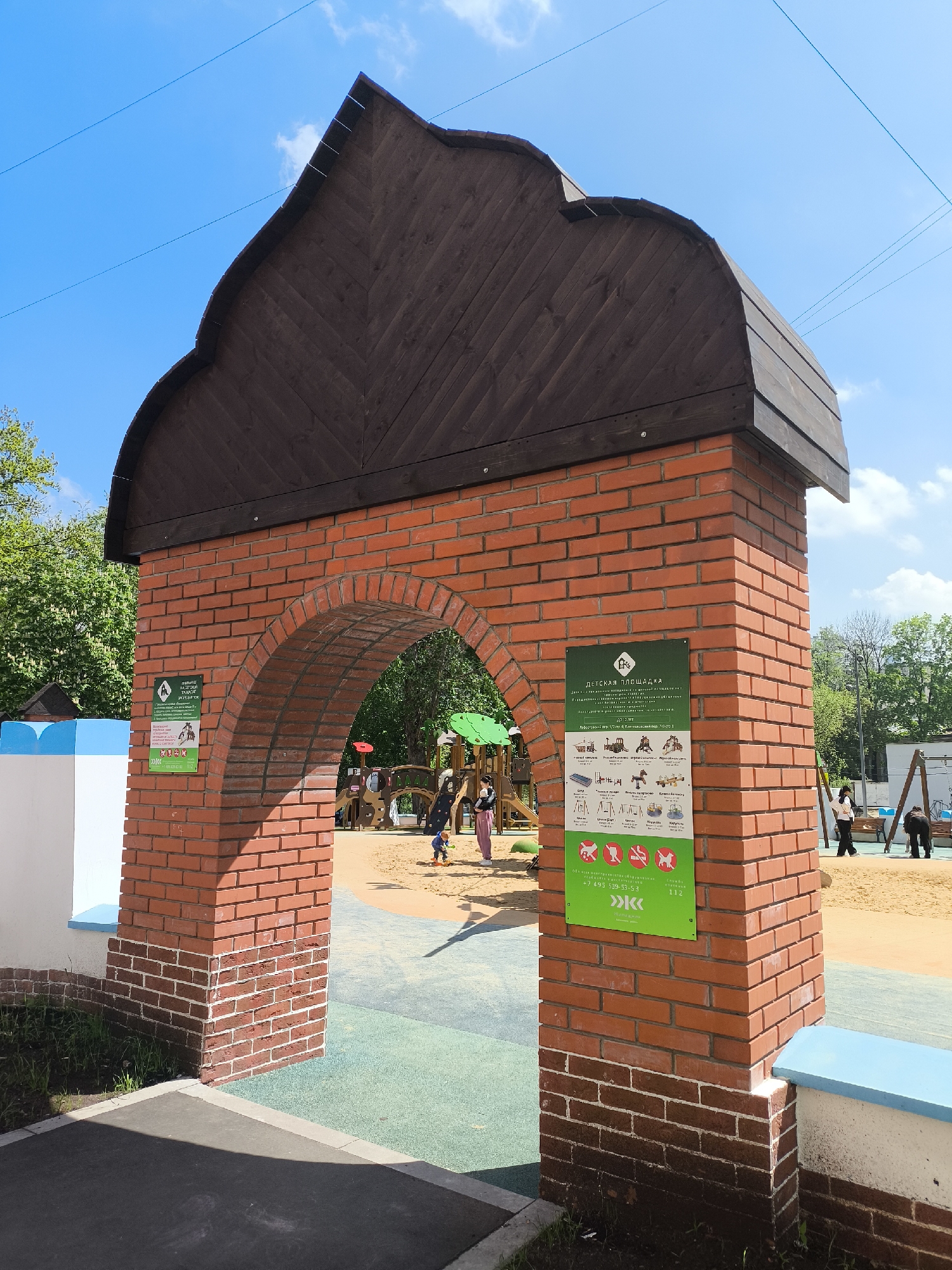 Here's a detailed description of the image:
**Overall Impression:**
The image captures the entrance to a playground, marked by a brick archway topped with a dark wooden roof. The setting appears to be a public park or recreational area.
**Subjects and Objects:**
* **Archway:** The most prominent feature is the brick archway. The arch is constructed of red bricks, forming a rounded opening. The top of the archway is capped with a dark brown wooden roof, slightly arched.
* **Signage:** Two signs are attached to the brick pillars of the archway. The sign on the right side of the picture displays the words "ДЕТСКАЯ ПЛОЩАДКА" with details in Cyrillic. The sign on the left side of the picture also displays details in Cyrillic.
* **Playground:** Through the archway, various elements of a playground are visible, including a sandpit, play structures (possibly climbing frames), and swings. People, mainly children and adults, are present within the playground area.
* **Background:** The background features trees and greenery, suggesting a park-like environment.
* **Pavement:** A section of pavement is visible in the foreground, near the base of the archway.
* **City Details:** The name of the city is Unknown, Unknown. The sign has details in Cyrillic indicating the exact location in the city.
**Environment:**
* **Weather:** The image was likely taken on a clear day with blue skies.
**Actions and Interactions:**
* People can be seen walking and playing in the playground, implying activity and usage of the park facilities.
**Forensic Details:**
* **Construction Materials:** The archway is primarily constructed of bricks and wood.
* **Signage:** The signage provides location details of the playground, details in Cyrillic.
* **State of Repair:** The structure seems to be well-maintained, with no visible signs of significant damage.