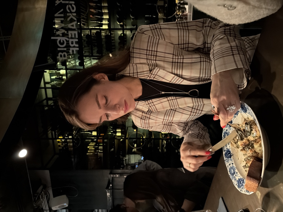 A woman with long brown hair is seated at a restaurant table, wearing a tan and white plaid blazer. She is holding a fork and knife, eating food from a blue and white patterned plate. The table is dark and there are other people sitting nearby. In the background, a wine rack filled with bottles is visible. The lighting is dim, creating a somewhat intimate atmosphere.