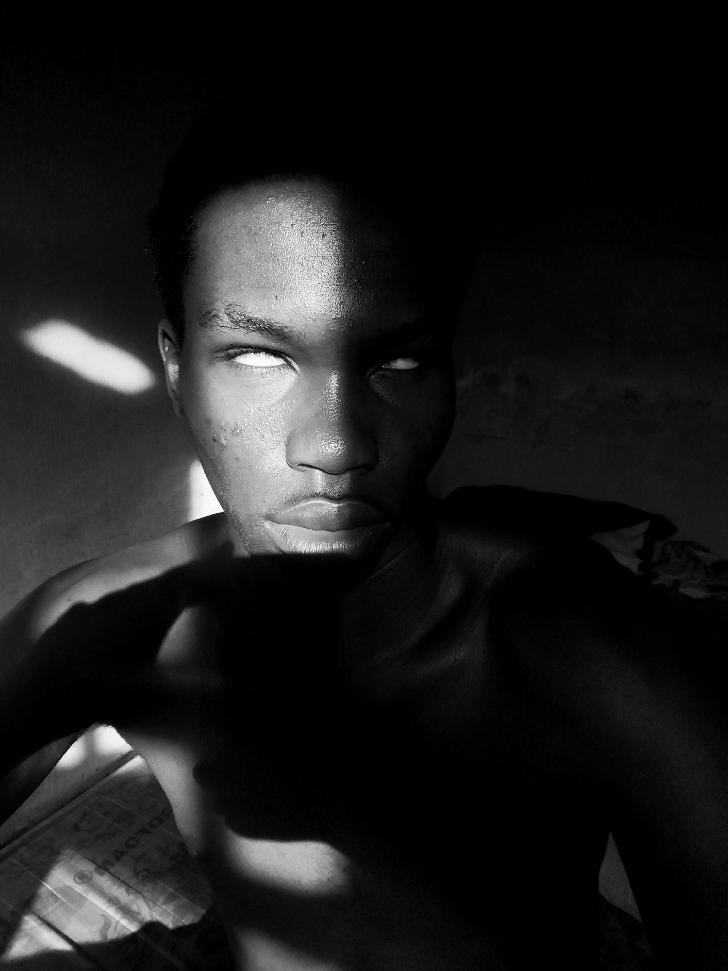 The image is a black and white close-up of a dark-skinned individual with their eyes rolled back, revealing the whites. A stark line of light illuminates the right side of their face, contrasting sharply with the darkness enveloping the rest. The person is shirtless, and a shadow obscures the lower part of their torso. The background is dark and indistinct. There is no verifiable information to place the individual in Keffi, Nigeria, or elsewhere.