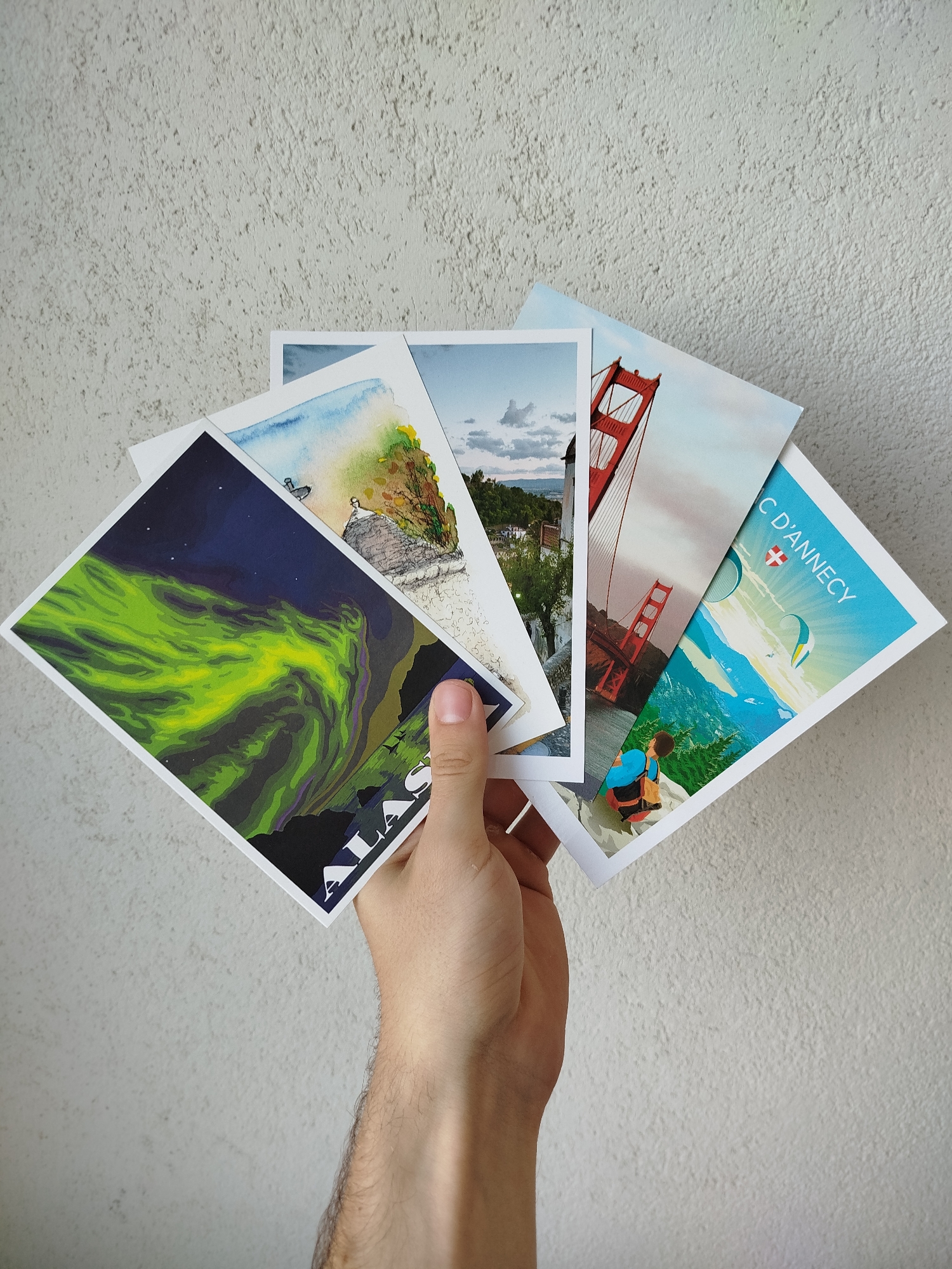 A human hand holds a fan of five postcards against a textured, off-white wall. The postcards vary in imagery and theme. The front postcard features a turquoise design with the words "C D'ANNECY" and a Swiss flag emblem. The second postcard shows a portion of the Golden Gate Bridge in San Francisco. The third has an urban view. The fourth is a watercolor painting of a coastal scene with a seagull. The fifth and furthest forward is a depiction of the Aurora Borealis with "ALASKA" printed on the bottom. The cards are slightly overlapping.