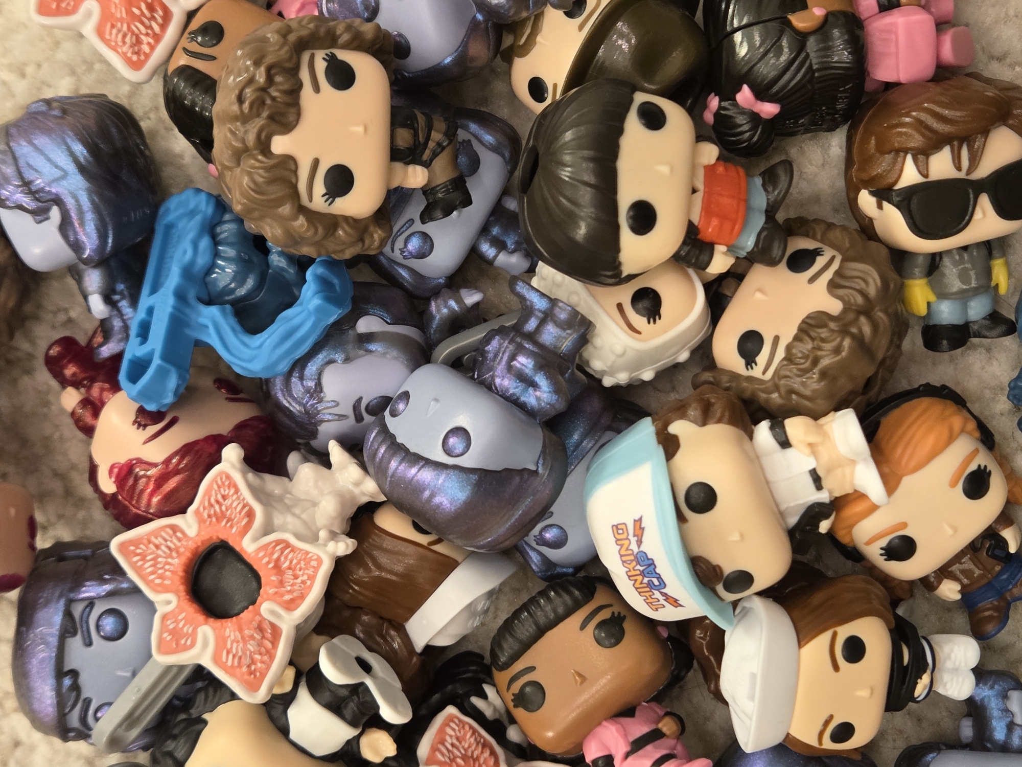 The image shows a close-up of a pile of Funko Pop figures. The figures are from the television show Stranger Things and are various characters. The figures include Eleven, Dustin, Steve, and a Demogorgon. The location is not included in the image.