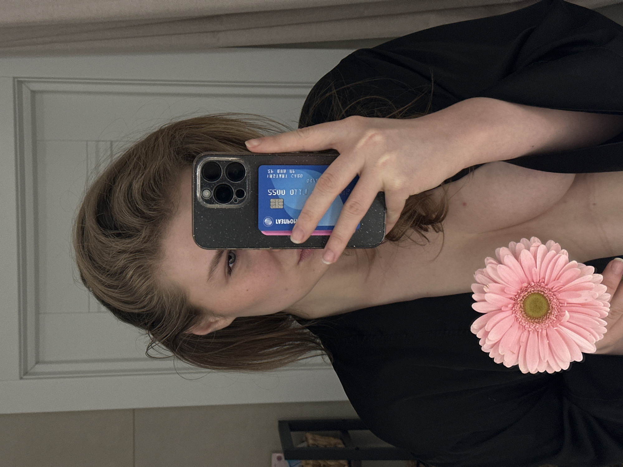 The scene depicts a female individual taking a mirror selfie indoors in Kazan, Russia. The subject has light brown hair and is partially visible, showing her left eye, nose, cheek, and chin, looking towards the camera's reflection. She is wearing an open black robe, revealing her bare chest and upper torso. Her right hand holds a dark-colored smartphone, consistent with an iPhone Pro model, in a clear, speckled case. A blue and pink bank card, with the text "МОРНЕАТ" (MORNAT), "5500 0113", a "MIR" payment system logo, and an expiration date of 08/25, is positioned between the phone and its case. Her left hand holds a single pink gerbera daisy flower with a prominent green center at mid-chest level. The background features a light-colored, paneled door behind the subject, with a dark, open-shelf unit partially visible to the left and a light-colored curtain or wall to the right. The environment appears to be a private domestic space.