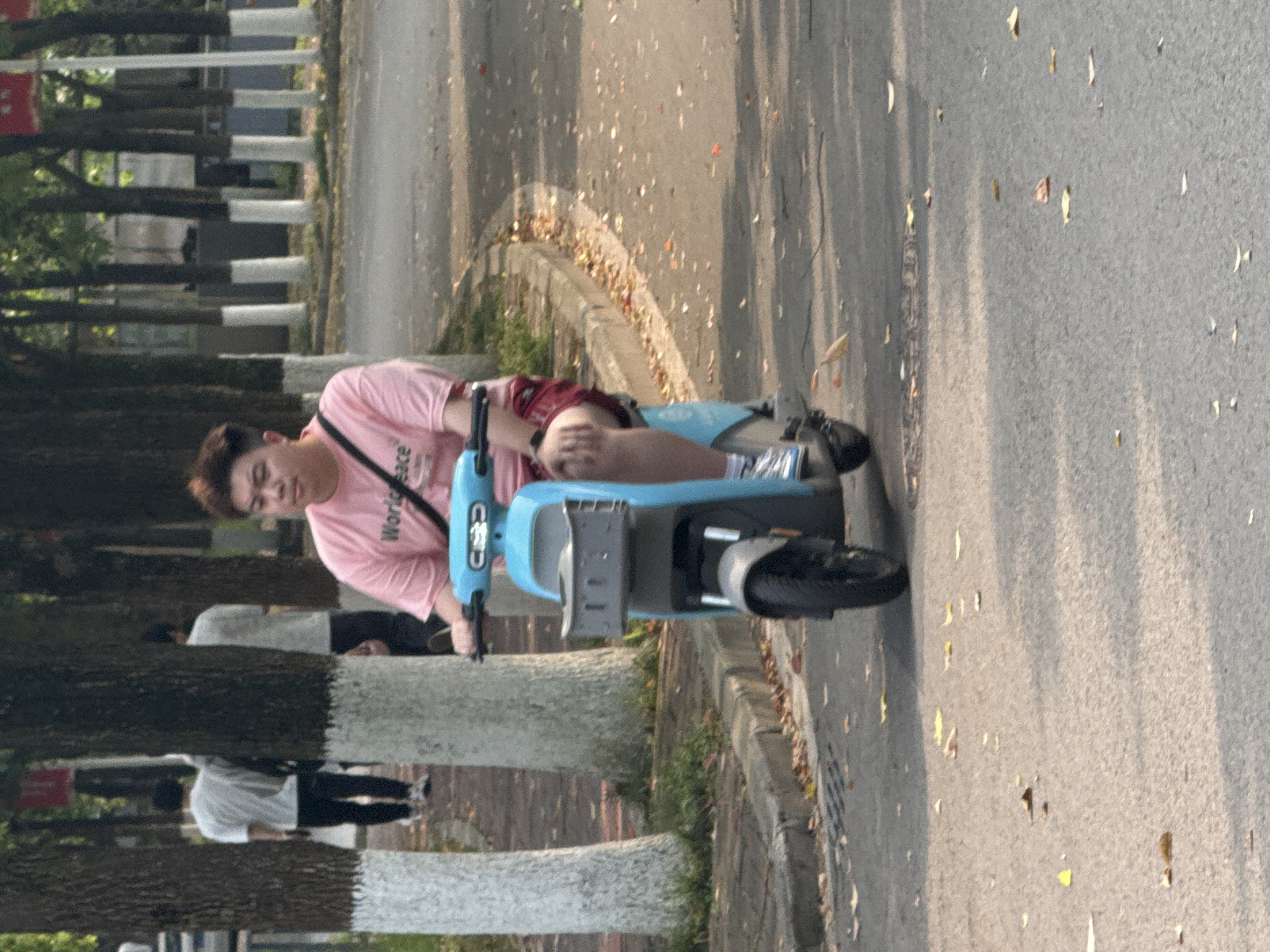 A man is riding a light blue electric scooter on a paved road. He is wearing a pink t-shirt with the words "World Peace" on the front, red shorts, and white shoes with socks. A black strap runs diagonally across his chest. The road is littered with fallen leaves, and the lighting suggests it's daytime.
Along the left edge of the image, there are trees with black and white striped trunks. In the background, a person wearing a white shirt and black pants is walking away from the camera. The road is likely a public thoroughfare in Jiangjin, China, based on the style of the scooter.
