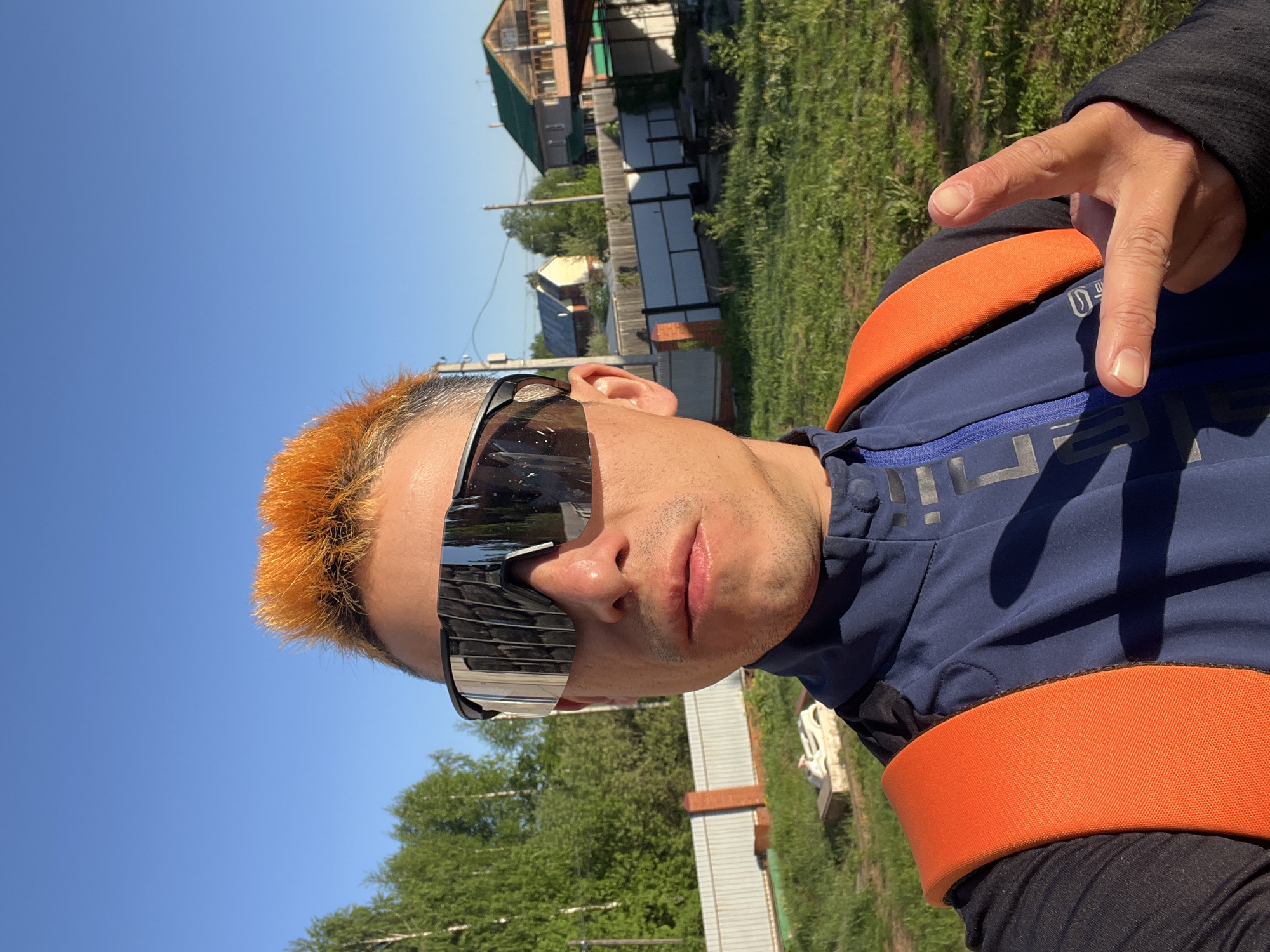 A man is posing for a selfie in an outdoor setting. He is wearing sunglasses and a blue and orange athletic jacket. His hair is dyed orange. He is holding up his right hand with his index finger and thumb extended. The background includes a blue sky, green trees, a house, and grass. The location is Chaikovsky, Russia.