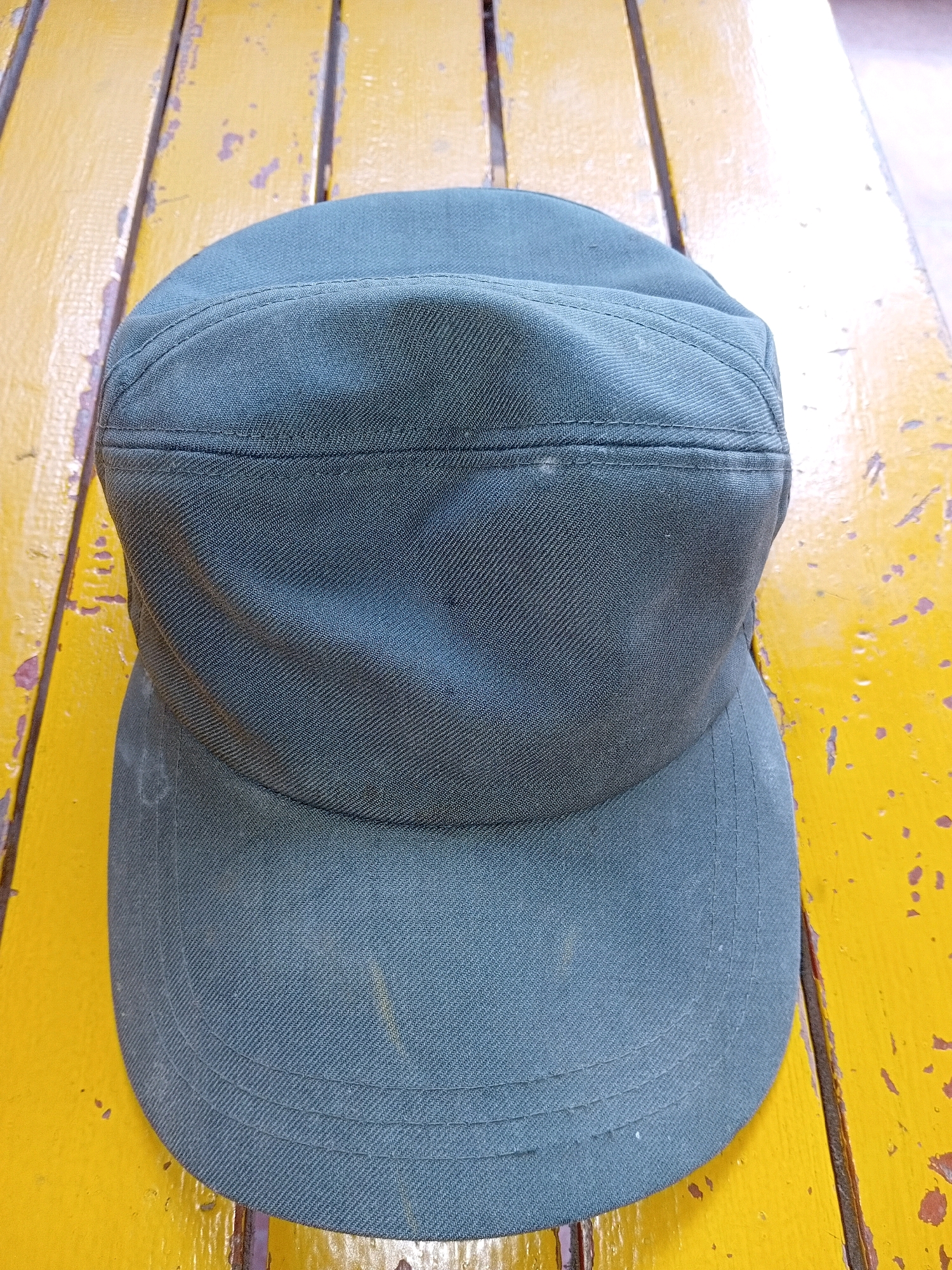 A dark green cap rests on a weathered, bright yellow wooden surface. The surface appears to be a table or bench made of planks, with visible wear and tear, including peeling paint and a grainy texture. The cap is positioned centrally in the frame, with its brim facing towards the viewer. The fabric of the cap has a visible weave, and there are stitched details on the crown and brim. The lighting suggests an outdoor or well-lit indoor environment, casting subtle shadows. The image does not contain any people or indicate a specific location within Aditirto, Indonesia.