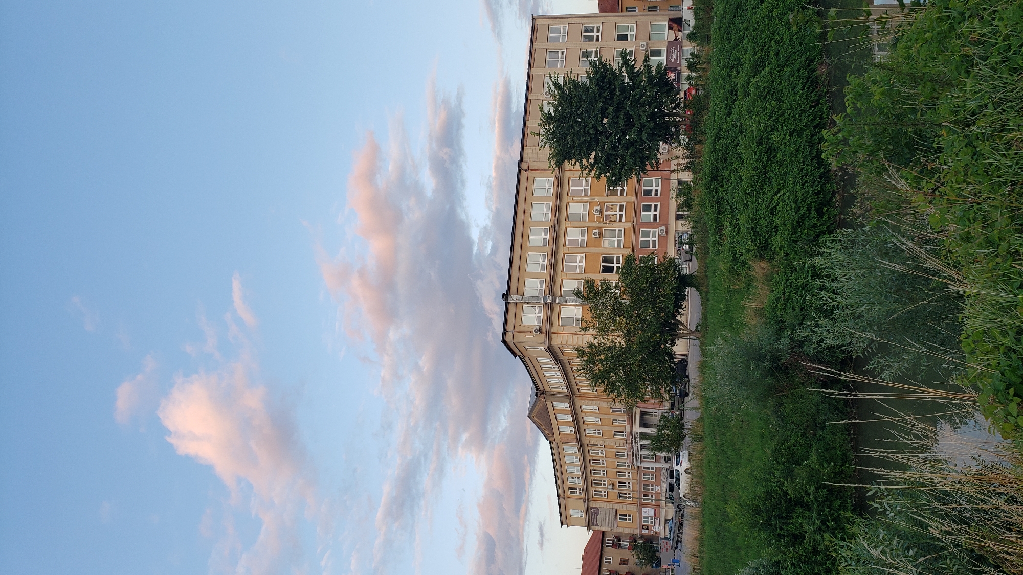 The image depicts a multi-story building with a yellow facade, situated in the city of Prešov, Slovakia. The building features numerous windows with white frames. In front of the building, there is a green grassy area with trees and some parked cars. A body of water with tall grass is visible in the foreground. The sky is partly cloudy with soft pink and blue hues, suggesting it might be sunset. The overall scene is tranquil and captures an urban landscape with natural elements.