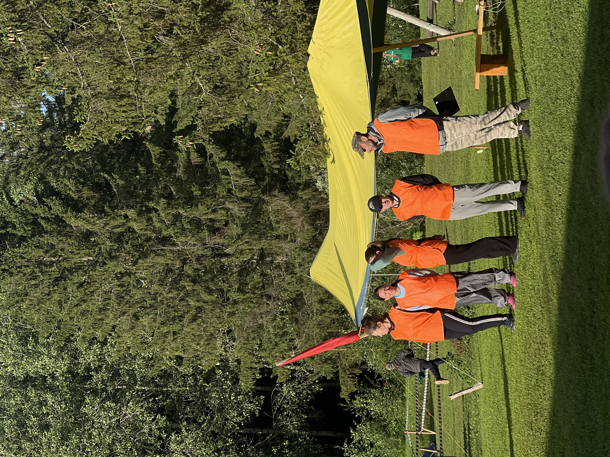 The image shows a group of six people standing in a line on a grassy area, with a yellow tent and a forest in the background. The people are all wearing orange vests over their clothing, suggesting they are participating in an organized activity or event. Some of them are wearing hats. The sun is shining, indicating it is daytime, and the presence of green grass and trees suggests a warm weather season, likely summer. The forest appears to be dense with pine trees. One person in the back is wearing a green shirt. The overall mood appears to be casual and perhaps focused on the activity. There is a red object that appears to be a flag or banner attached to the tent, and some wooden structures with ropes are visible in the foreground, hinting at an outdoor recreational or training course. No text is clearly visible in the image.