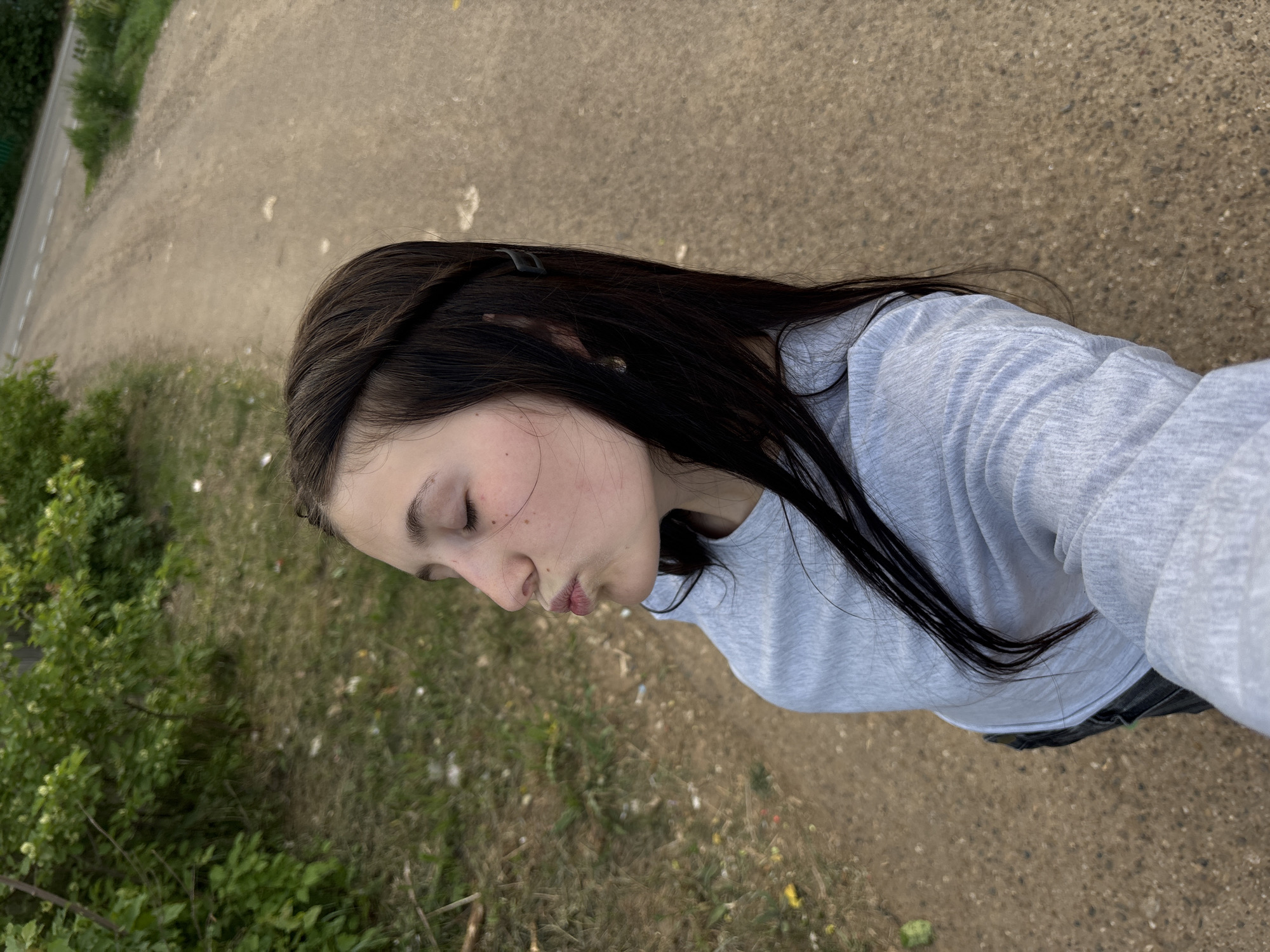 A young woman is captured in a close-up shot outdoors. She has dark brown hair with a hair clip, and is wearing a light gray long-sleeved shirt. Her eyes are closed. The background includes a path or road, as well as greenery, suggesting a park or roadside environment. No specific details indicate the exact location within Фоки, Russia.