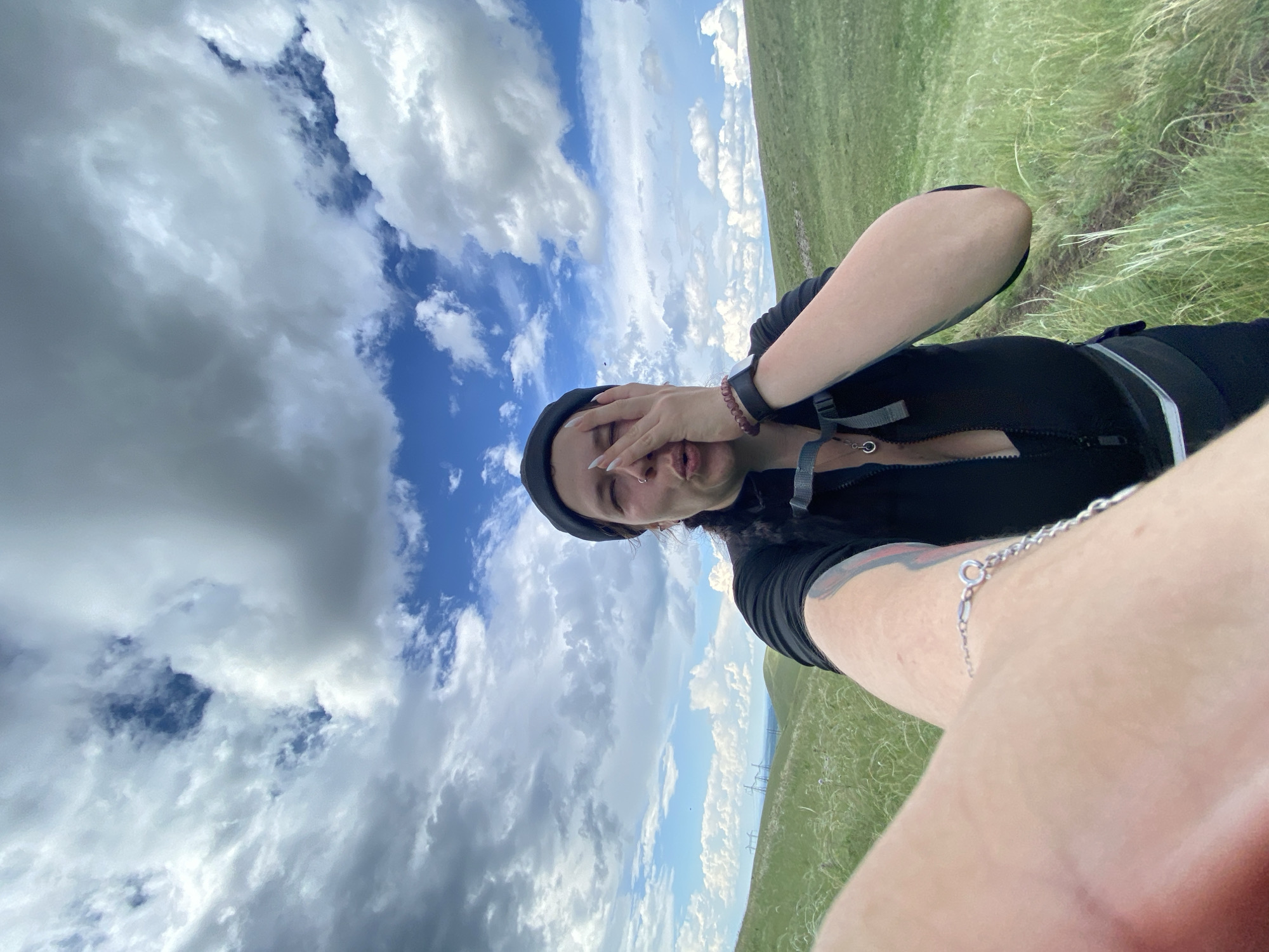 A woman is taking a selfie outdoors in a hilly, grassy area with a cloudy sky. She is wearing a black beanie, a black zip-up jacket over a black top, and dark pants. She has a silver bracelet on her left wrist and a black watch on her right wrist. Her face is partially obscured by her left hand, and she appears to be puckering her lips. The background shows rolling green hills with some power lines visible in the distance. The location is the city of Verkhneozernoe, Russia.