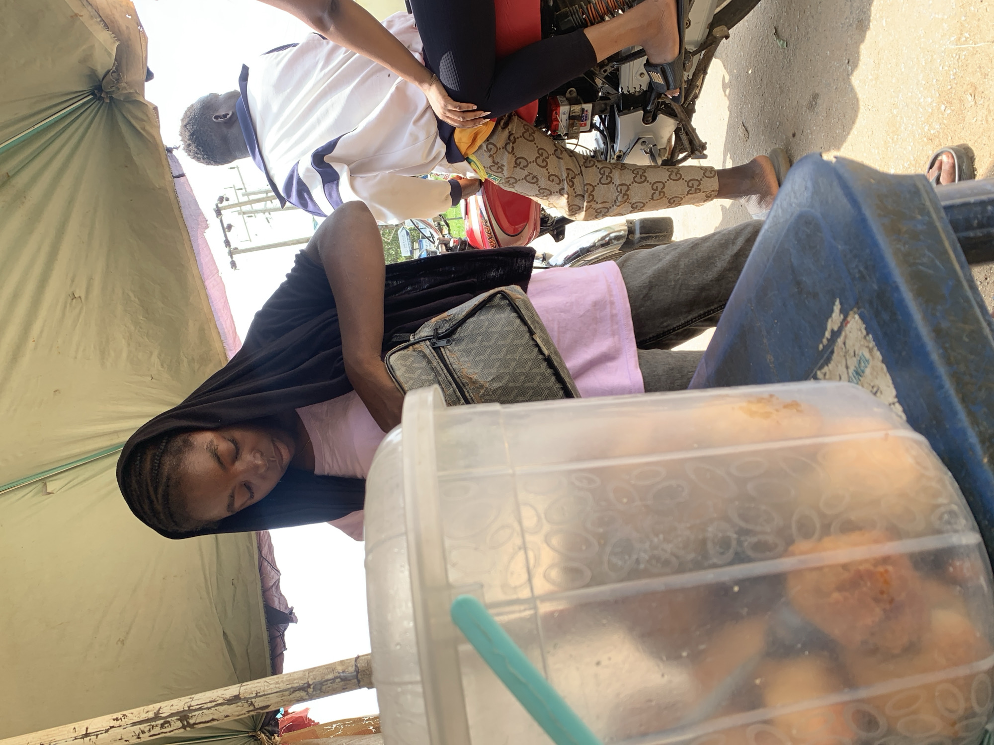 This media file shows a group of people in an outdoor setting, likely a street market or roadside stall in Abuja, Nigeria. The scene is oriented sideways. In the foreground, a person with braids, wearing a pink top and a black headscarf, is leaning back and appears to be resting or sleeping. A clear plastic container filled with what looks like baked goods, possibly buns or pastries, is prominently displayed in front of them, with a blue straw visible inside.
Behind this person, a motorcycle is partially visible. A second person, wearing a white jacket with blue stripes and patterned pants, is seated on the motorcycle. Their legs and feet, wearing sandals, are also visible. A third person's legs and feet are also visible, wearing black leggings and sandals, suggesting they might be on the back of the motorcycle or standing nearby.
The background shows a makeshift tent or shelter with a light-colored tarp, suggesting a vendor's stall. Overhead wires and utility poles are visible in the distance, indicating an urban or semi-urban environment. The lighting suggests it is daytime, with bright sunlight casting shadows. The overall impression is of a casual, everyday scene at a market or roadside business. No discernible text is visible in the image.
