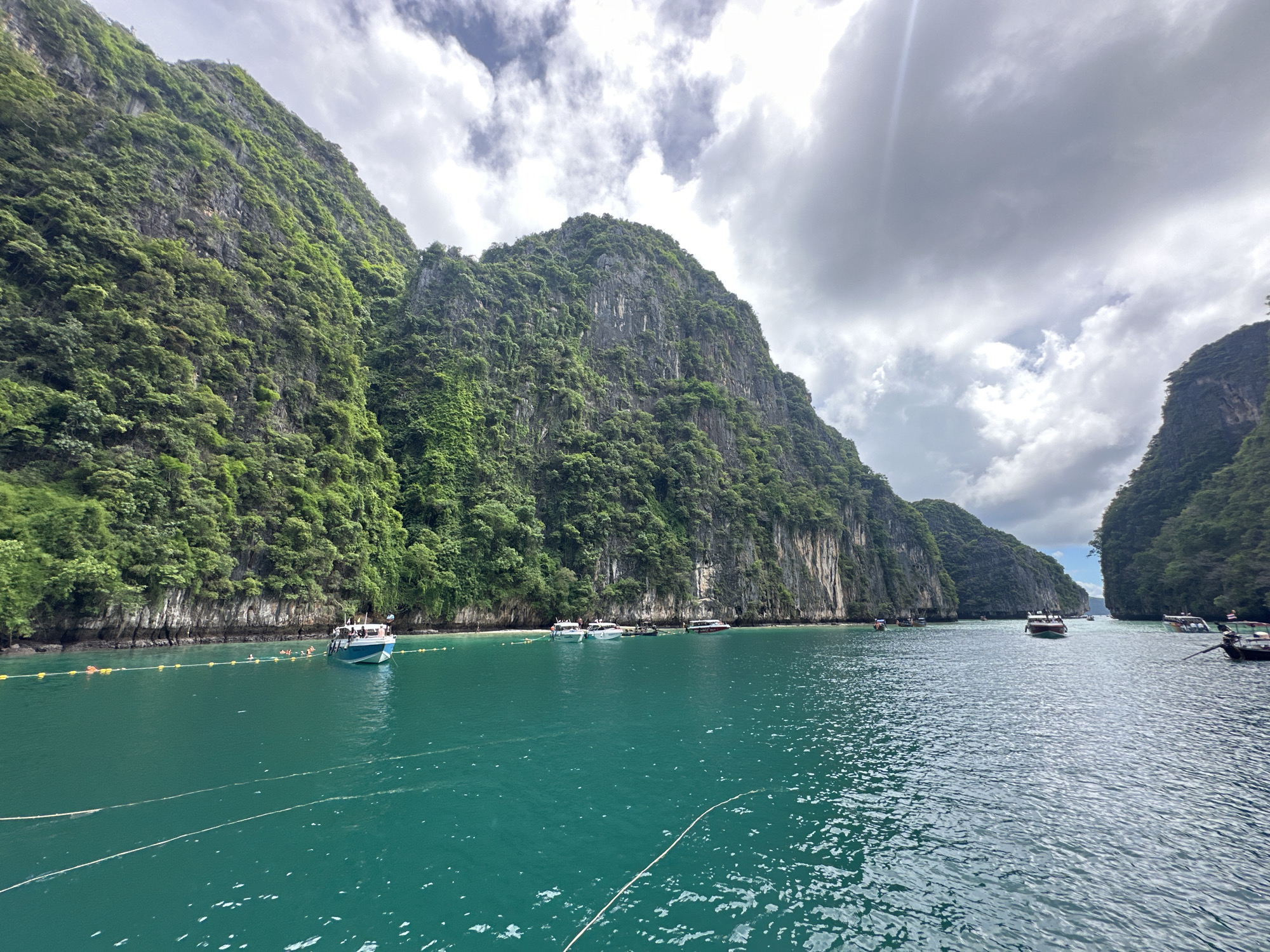 The image depicts a body of water with steep, tree-covered limestone cliffs rising on either side. The water is a vibrant turquoise color, and several boats are visible on the surface. In the distance, more cliffs and boats can be seen, suggesting a bay or cove. The sky is partly cloudy, with patches of sunlight breaking through. On the left side of the image, a group of people can be seen near a small boat anchored close to the shore, with yellow buoys and ropes marking an area in the water. The overall scene is one of natural beauty and recreational activity. The location is likely part of the Phi Phi Islands, which are a popular destination accessible from Ao Nang, Thailand.