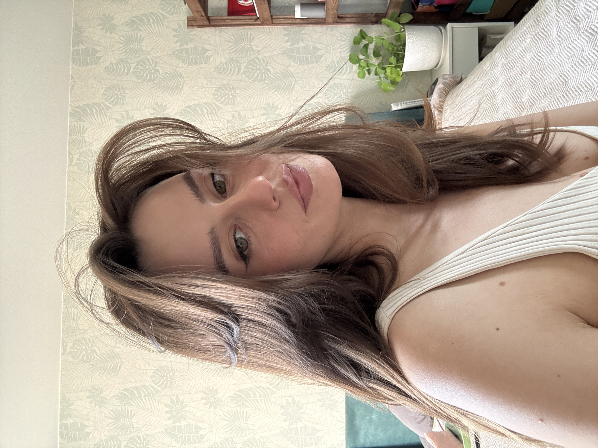 A head-and-shoulder portrait depicts a young woman in an indoor setting, taken from an overhead perspective and rotated ninety degrees clockwise. She has long, wavy brown hair that cascades around her face and over her left shoulder. Her head is slightly tilted, and she looks directly at the viewer with light-colored eyes, displaying a neutral and calm expression. She is wearing a light-colored, ribbed tank top.
The background reveals a room, likely a bedroom or living space in Krasnoyarsk, Russia, brightly lit by natural light, suggesting it is daytime. A wall covered in light-toned wallpaper featuring a subtle leaf pattern is visible behind her. Above and behind her, a light wooden shelf holds various items, including a white potted plant with green foliage and a book with the Russian text "КАК ИЗОБРЕСТИ" (How to Invent) visible on its spine. To her right, part of a bed or sofa with a textured, light-colored blanket can be seen. Small moles are visible on her shoulder.