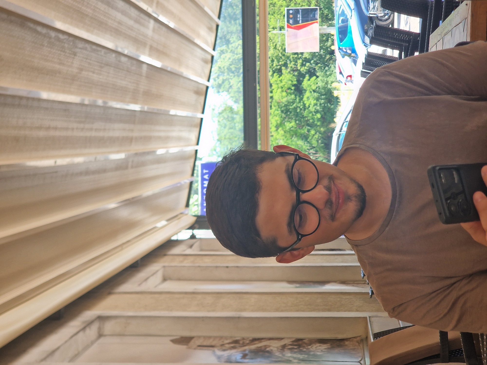 A young man with short dark hair, a goatee, and dark-rimmed glasses is looking down at his black smartphone, which he holds in his right hand. He is wearing a plain brown t-shirt. The man is seated at a table outdoors, with chairs and other tables visible behind him. The background suggests an urban environment with trees and parked cars. A building with light-colored columns and a retractable awning is to his left, and a blue sign is visible behind his head. This scene appears to be at a cafe or restaurant in the city of Cetinje, Montenegro, likely in a section with outdoor seating, possibly along one of the city's main boulevards like Njegoševa Street, given the presence of cafe culture and a visible street beyond the seating area.