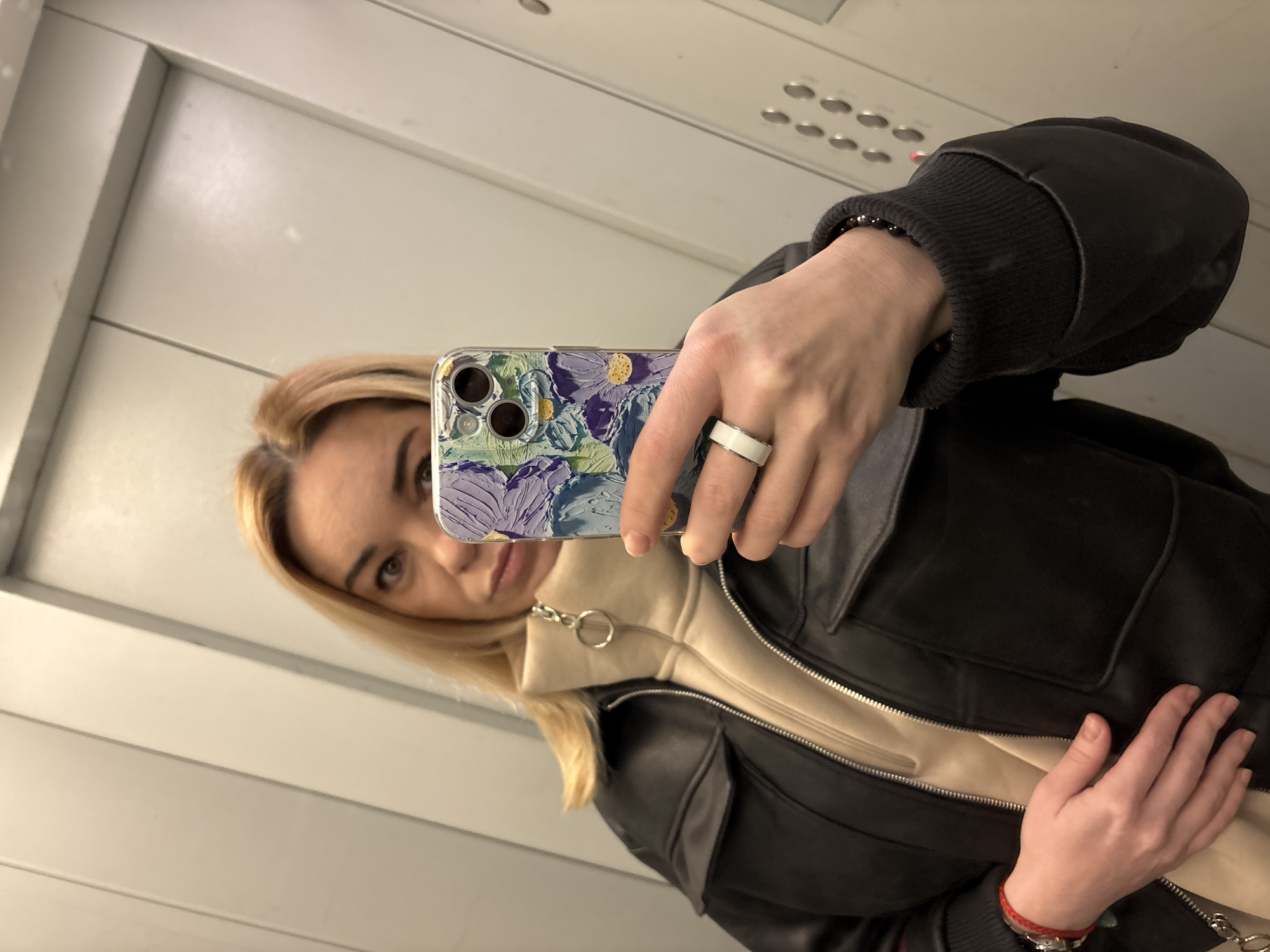 A woman with light blonde hair is taking a selfie in what appears to be an elevator. She is holding a phone with a floral patterned case in her right hand. She is wearing a black leather jacket over a beige zip-up sweatshirt and a white ring on her ring finger. On her left wrist, there is a red bracelet with possible embellishments. The elevator interior is plain with a white or light gray wall and ceiling. The location in Vsevolozhsk, Russia is unknown.