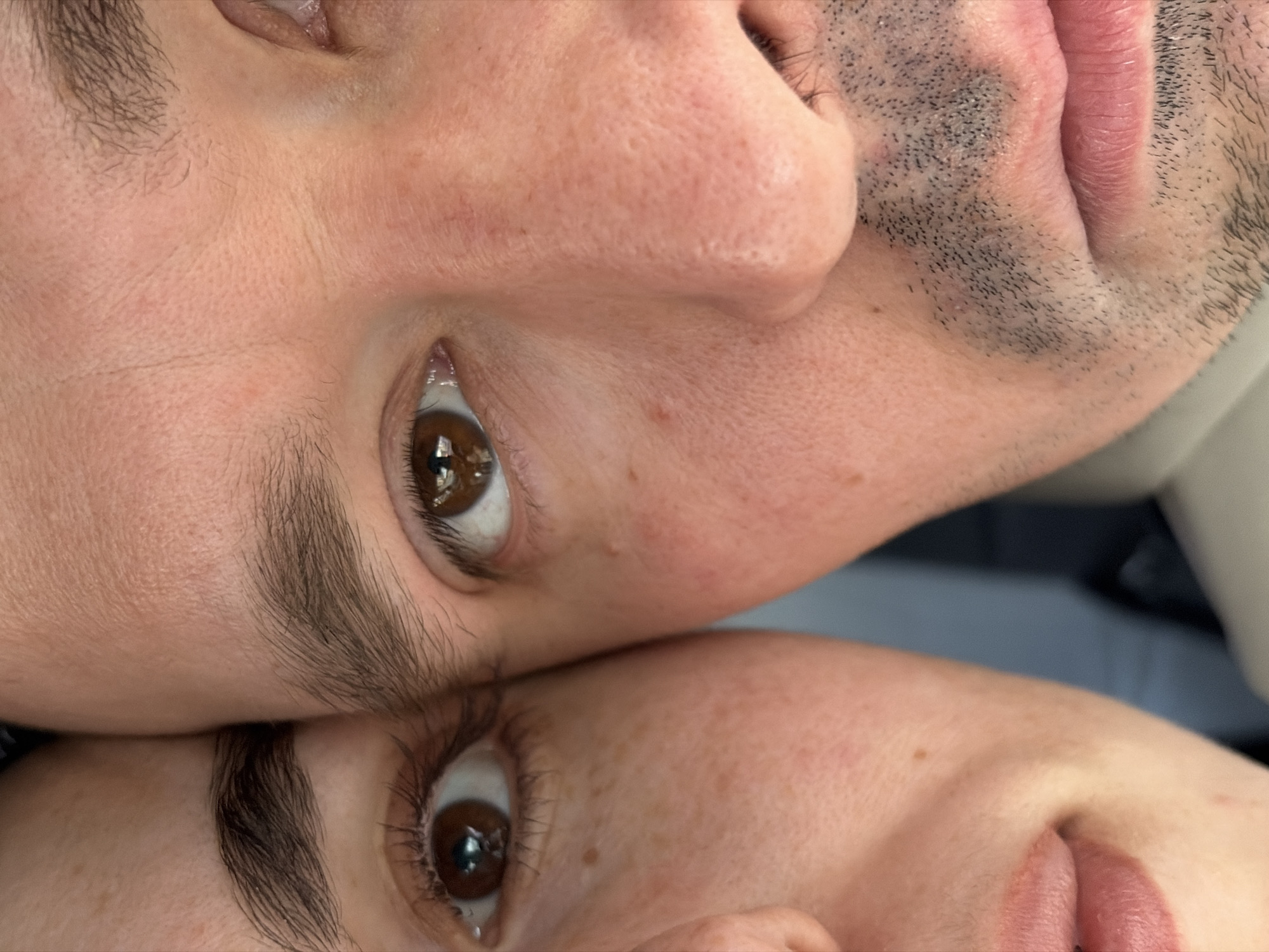 Two faces are closely positioned together in the image. The upper face appears to belong to a male, indicated by the presence of stubble or a beard. The lower face seems to belong to a female. Both individuals have brown eyes and are fair-skinned. The image does not depict any identifiable landmarks or clues to suggest the location is Izhevsk, Russia, or any other specific place. The background is out of focus, obscuring any environmental details. The primary focus is on the close proximity and facial features of the two individuals.