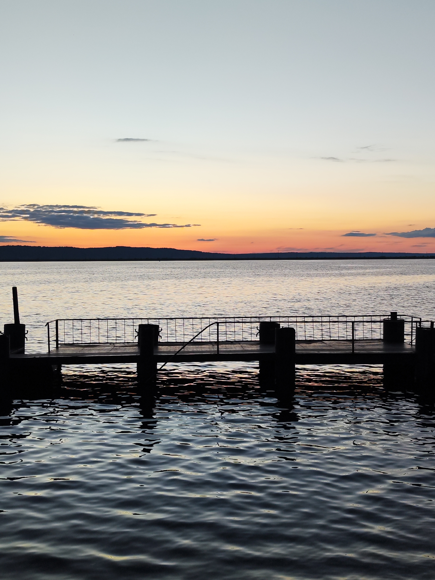 The image presents a serene twilight landscape over a large body of water, likely a lake or wide river, typical of a scene in Chaikovsky, Russia.
In the mid-ground, a small, dark-silhouetted pier extends into the water. The pier features a flat deck and a simple metal railing, supported by several thick posts that are partially submerged, creating subtle reflections on the water's surface.
The water occupies the lower two-thirds of the frame, displaying gentle ripples. In the immediate foreground, the water appears dark, almost black, reflecting very little light. As it stretches towards the horizon, it gradually lightens to a silvery-grey, reflecting the subdued sky.
A distinct horizon line separates the water from a distant, dark landmass, which appears as an undulating silhouette across the entire width of the image. Above this, the sky exhibits a striking gradient of colors indicative of either sunset or dusk. The upper part of the sky is a pale blue-grey, transitioning smoothly to soft yellows, and then to warm oranges and reds directly above the distant land. Scattered clouds are visible, including some small, wispy formations in the lighter upper sky and a few darker, more defined clouds closer to the horizon.
There are no people or specific activities depicted in the scene, conveying a sense of calm and solitude. No visible text is present in the image.
