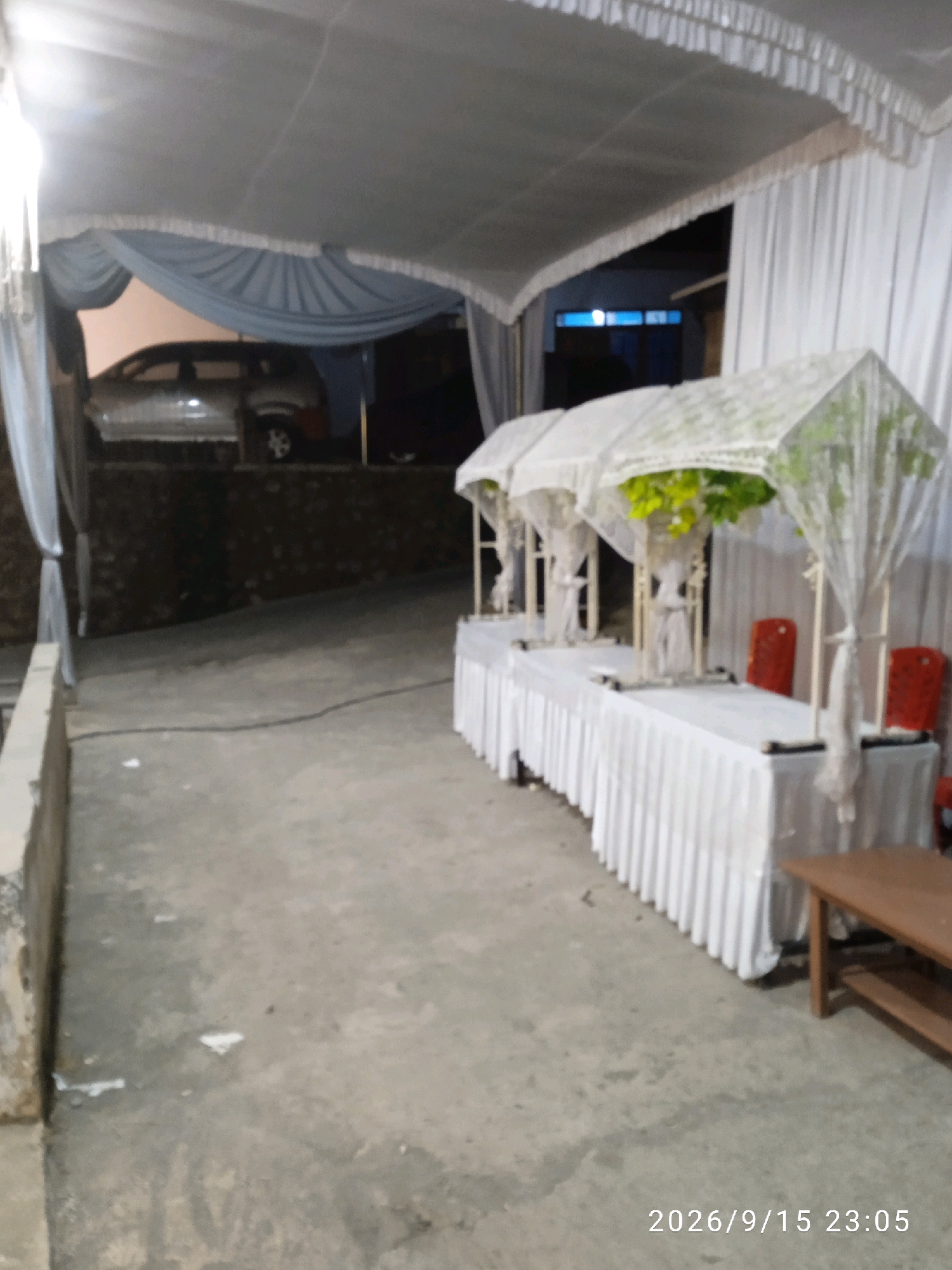 An outdoor, covered event setup is depicted, likely in Kelurahan, Indonesia, with a date and time stamp of "2026/9/15 23:05." The foreground shows a concrete pathway leading into the scene. To the right, a long, white-skirted table is set up with decorative elements. Three tiered umbrella-like structures, adorned with lace and faux greenery, are spaced along the table. Two red chairs are visible behind the table. In the middle ground, a car is parked on an elevated surface behind a retaining wall, partially obscured by a blue draped fabric. The background reveals a dark, possibly residential area, with some illuminated windows. The scene appears to be set up for an event, possibly a wedding or reception, given the draped decor and table setting. The overall lighting suggests it is nighttime.
