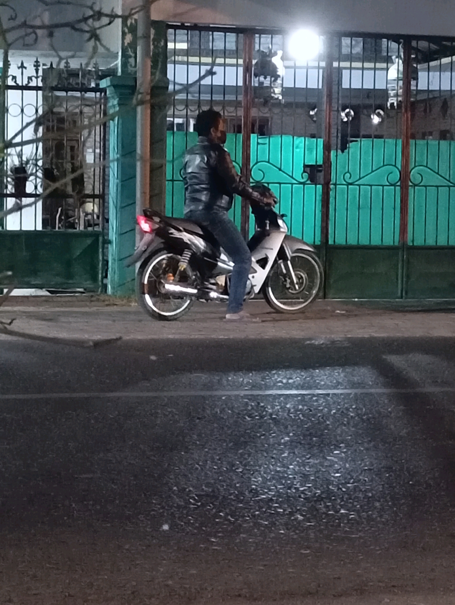 A person wearing a black jacket and blue jeans sits on a light-colored motorcycle in front of a green gate. The person has dark hair and is wearing a face mask. The motorcycle is parked on a sidewalk, with its front wheel on the right and its rear wheel on the left. The background shows a black metal fence and a light source reflecting on the ground. The scene appears to be outdoors at night, possibly in Randegan, Indonesia, given the context.
